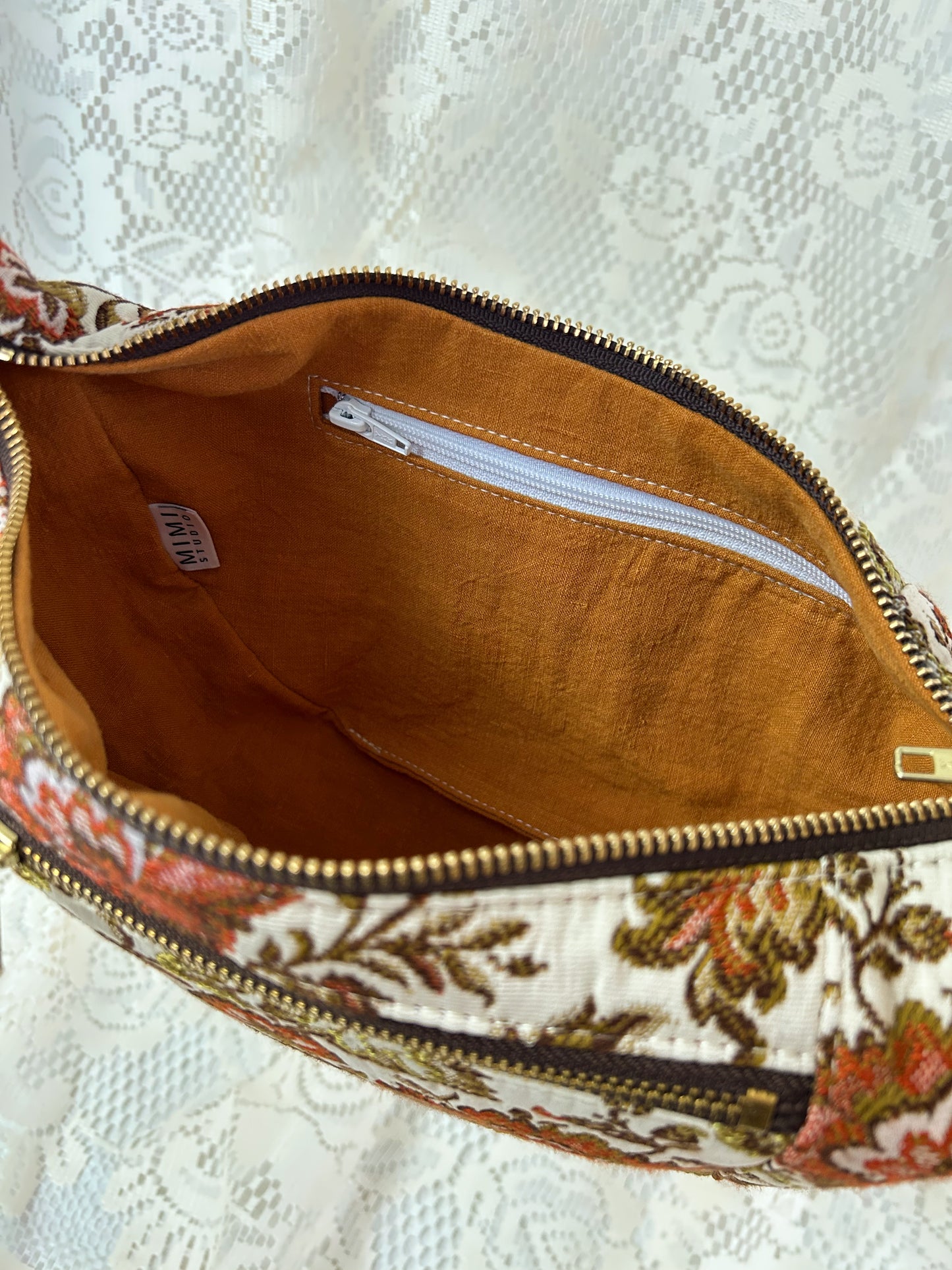 Close up of the lining fabric for the floral upholstery fabric crescent bag which is burnt orange linen and has a zippered pocket, displayed in front of a white lace backdrop.