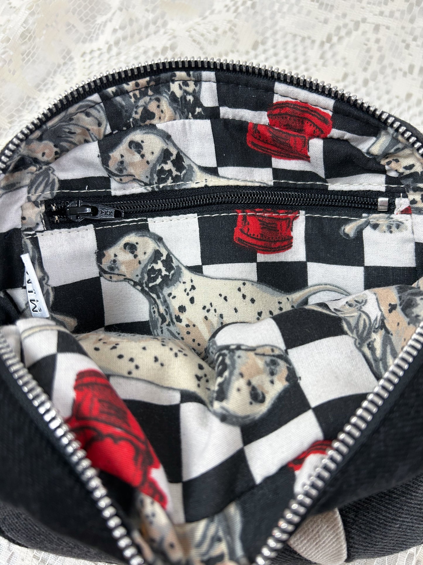 Lining fabric for the grey, black and ivory denim fanny pack with a patchwork star pattern which is a cotton-poly blend fabric with a dalmation and fire hydrant pattern and has a zippered pocket, on a white lace background