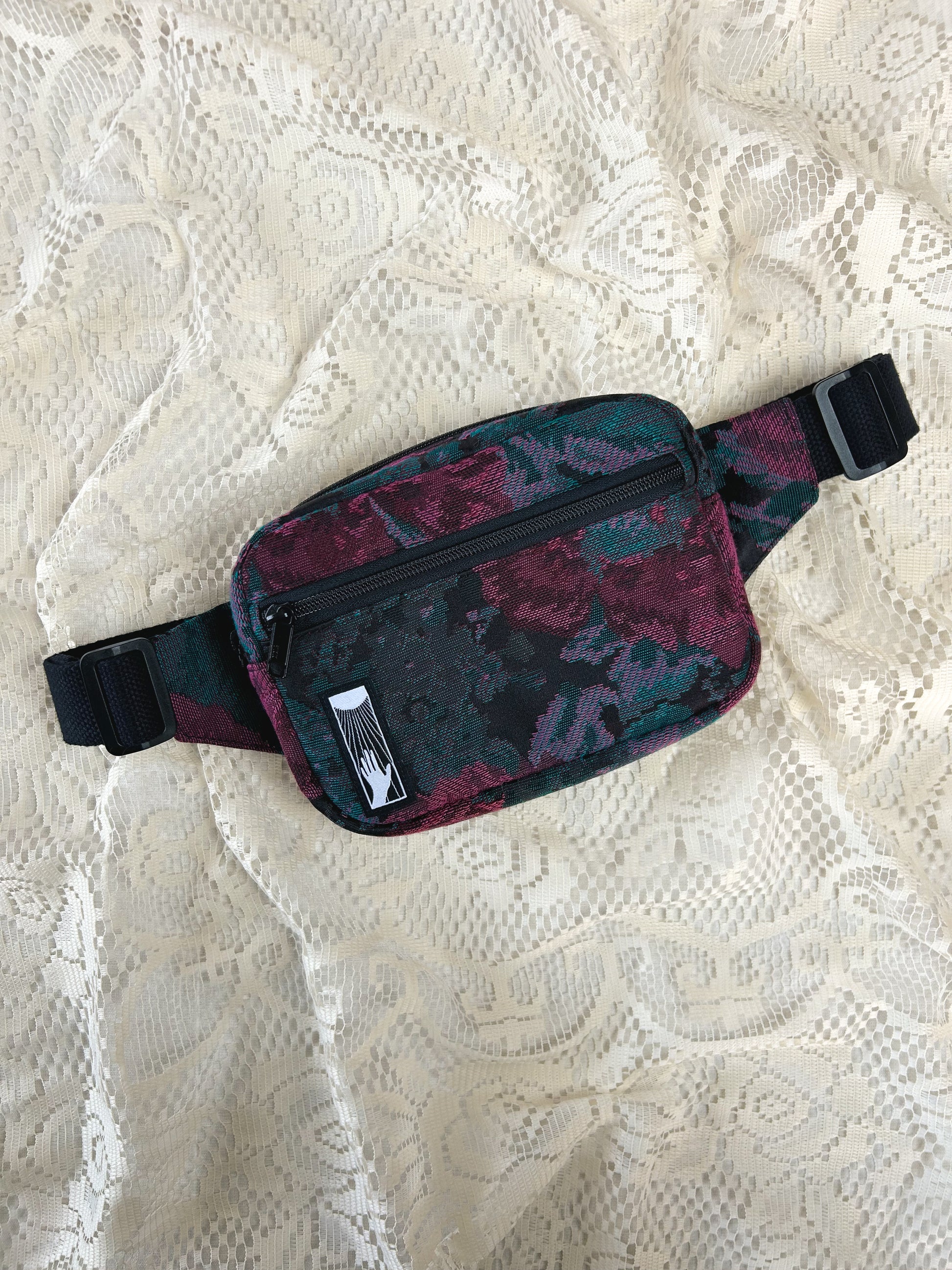 Flat lay view of the black, burgundy, and turquoise floral patterned upholstery fabric fanny pack with front zippered pocket on an ivory lace backdrop