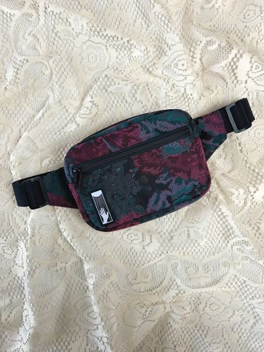 Flat lay view of the black, burgundy, and turquoise floral patterned upholstery fabric fanny pack with front zippered pocket on an ivory lace backdrop