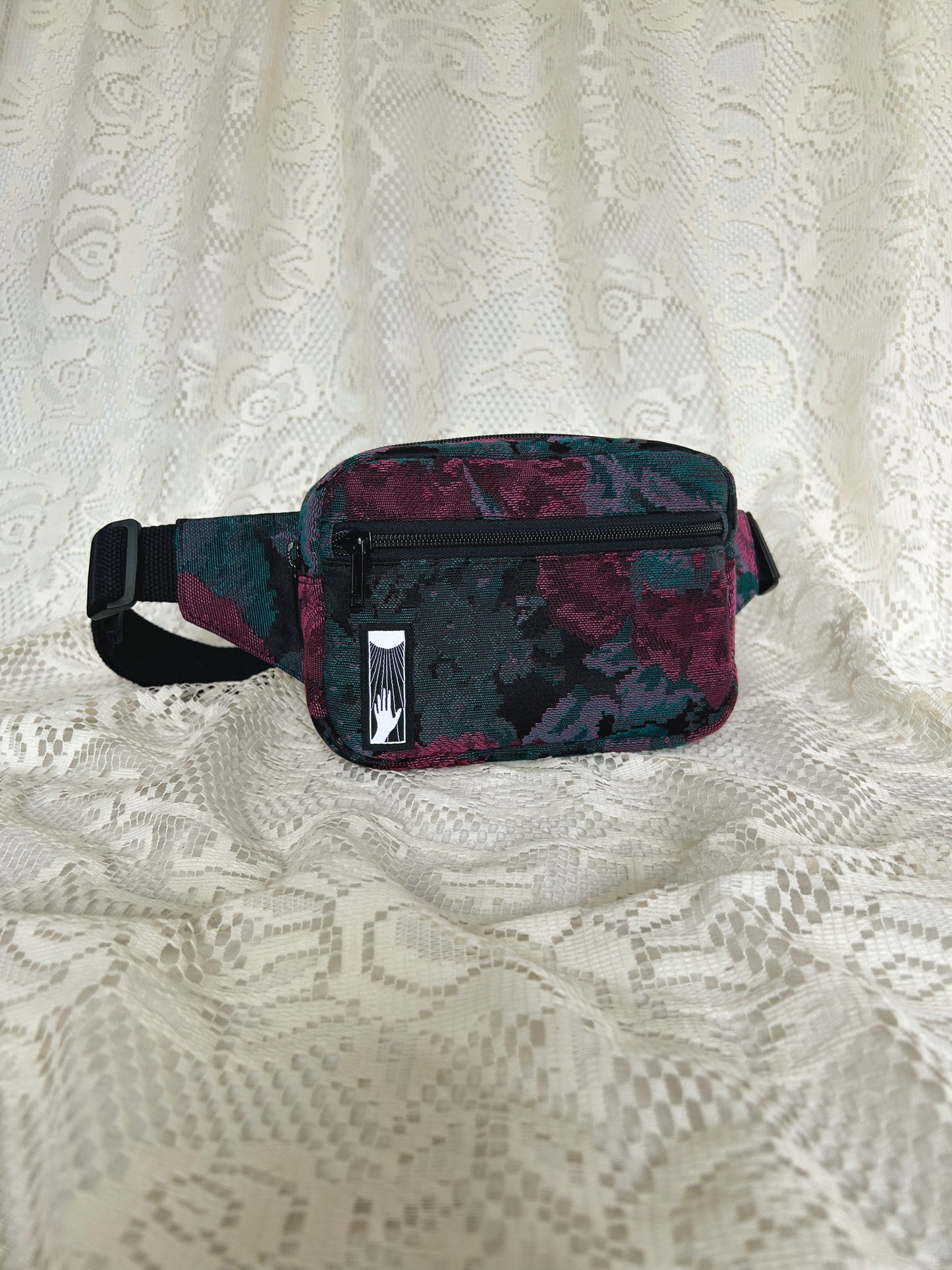 Angled front view of black, burgundy, and turquoise floral patterned upholstery fabric fanny pack with front zippered pocket with an ivory lace backdrop