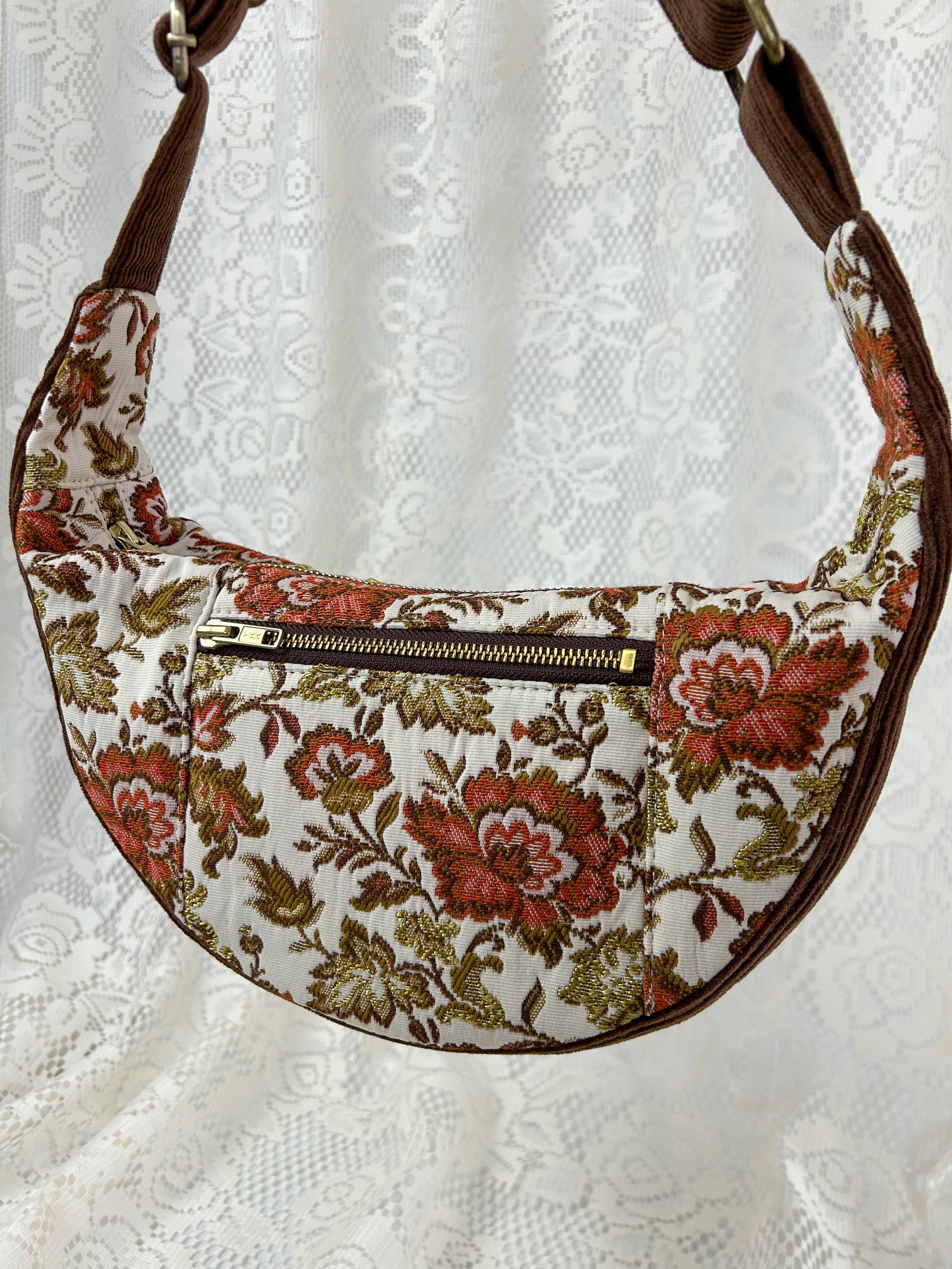 A second close up shot of the front zippered pocket on the floral upholstery crescent bag displayed in front of a white lace backdrop.