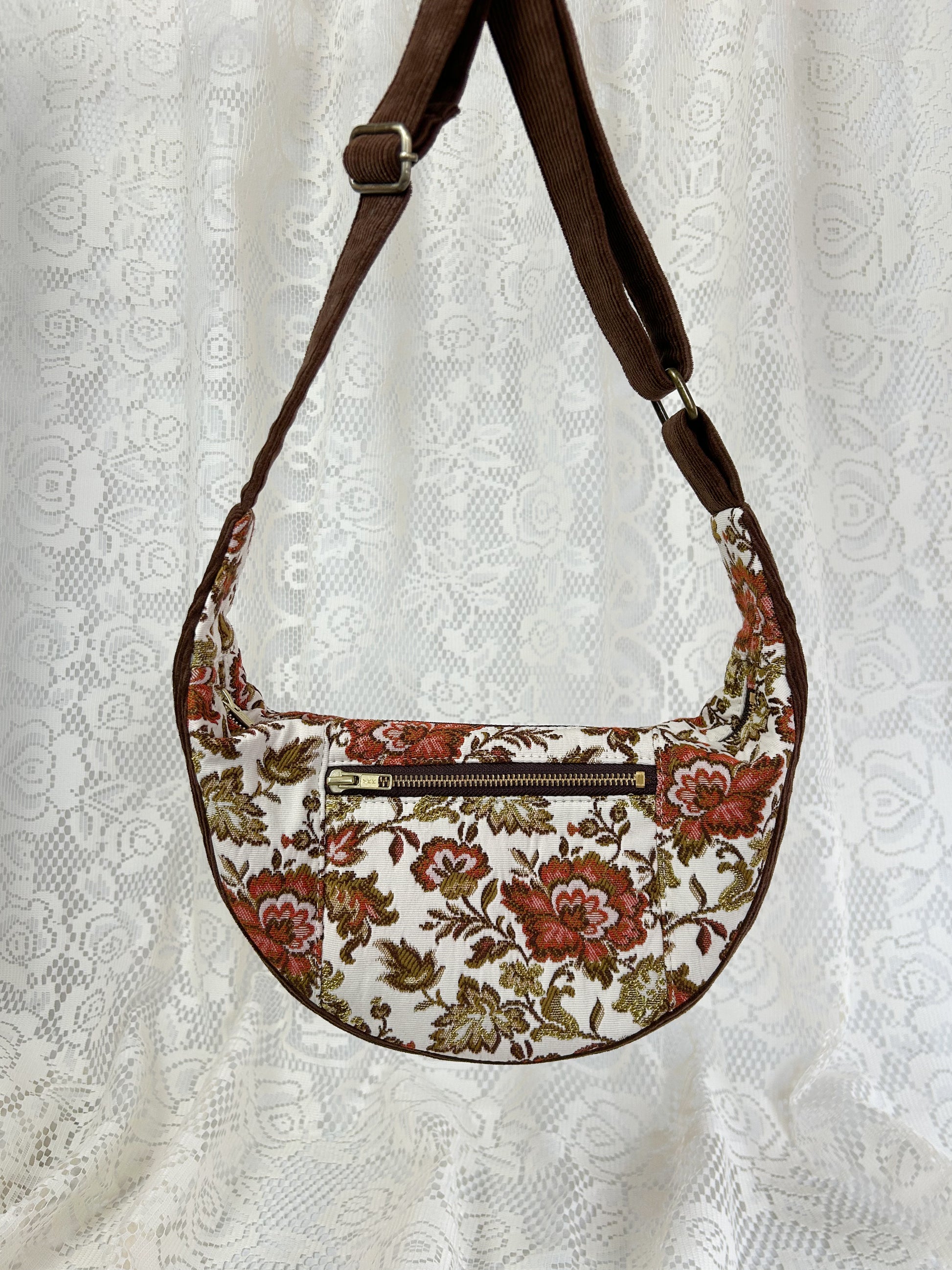 Floral upholstery fabric crescent bag with a brown corduroy strap in front of a white lace backdrop