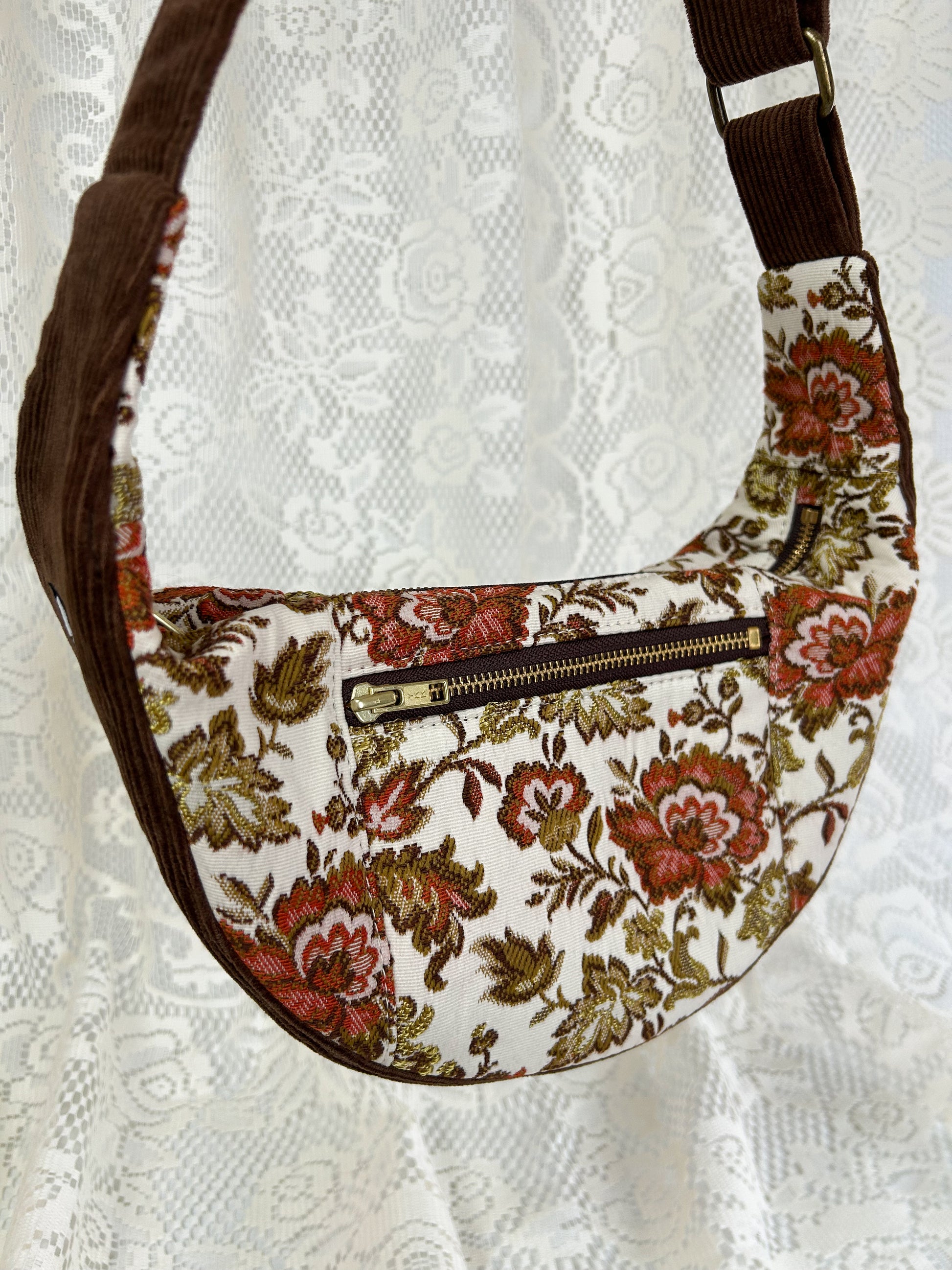 Close up of the front zippered pocket on the floral upholstery crescent bag displayed in front of a white lace backdrop.