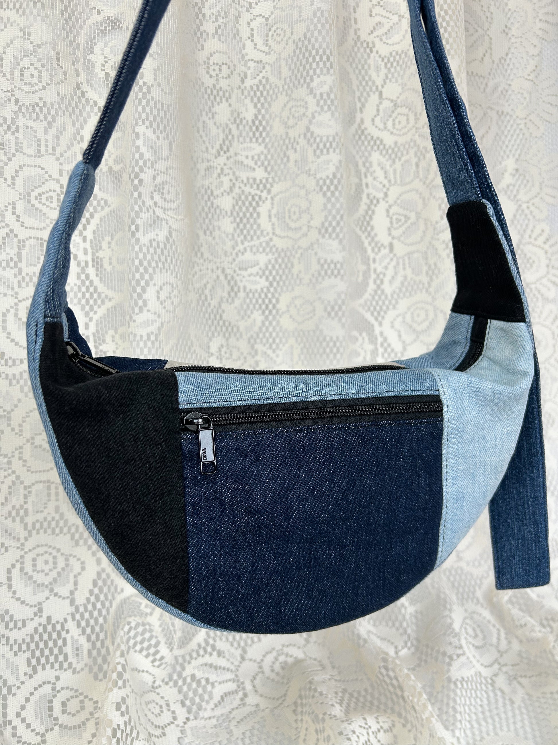 Close up of the multi-coloured denim crescent bag with a zippered pocket in front of a white lace backdrop
