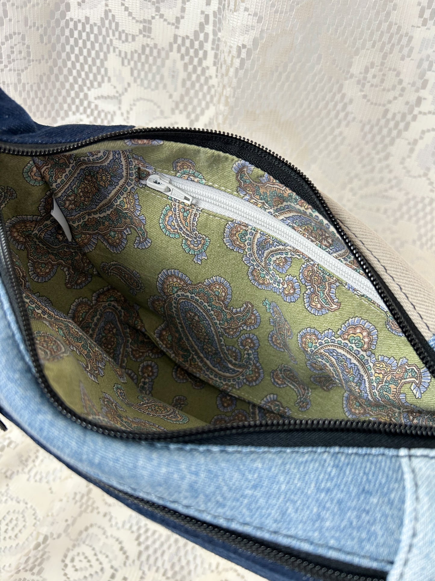 Close up of the lining for the multi-coloured denim crescent bag which is a paisley pattern cotton and has a zippered pocket, in front of a white lace backdrop