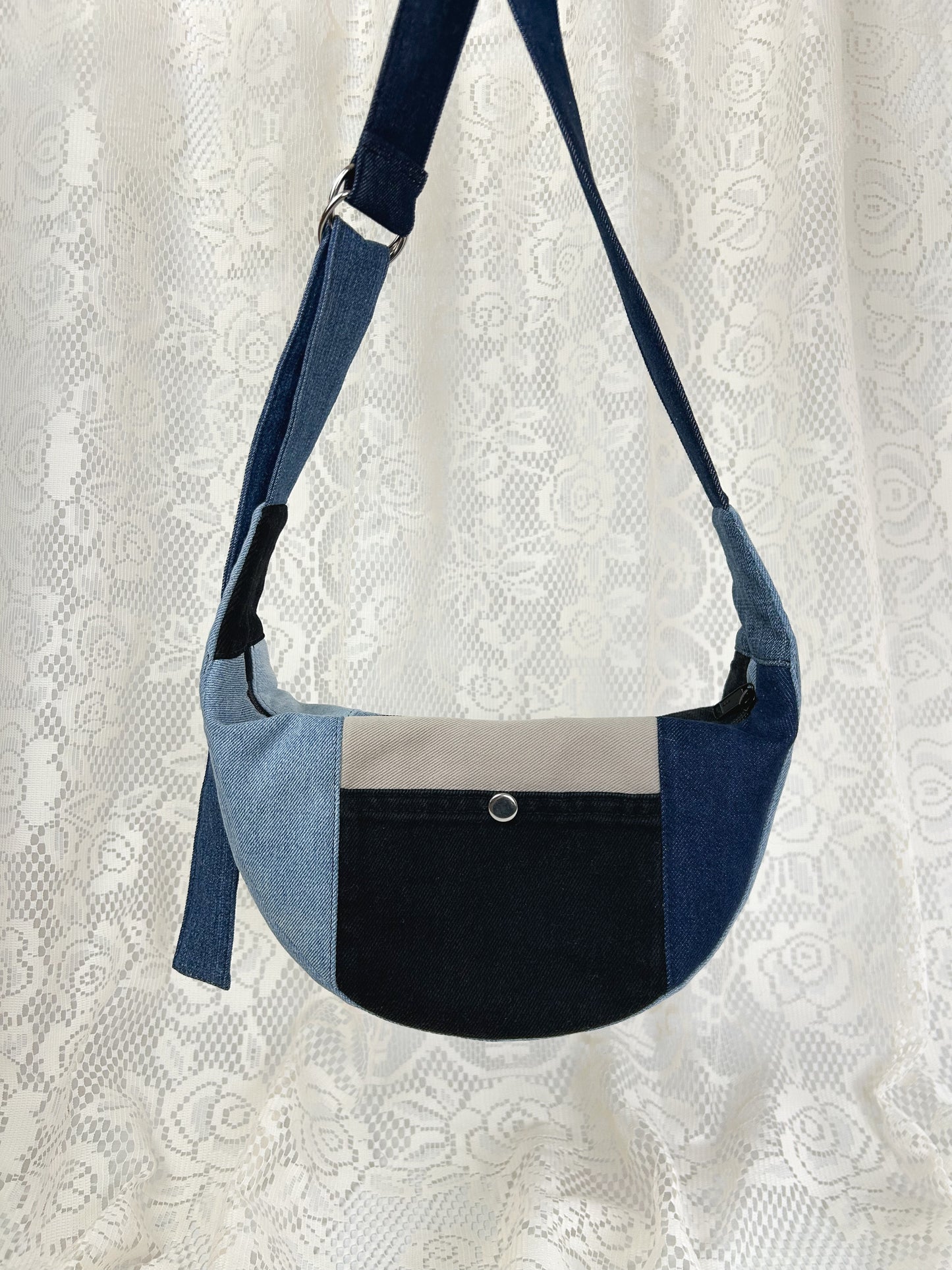Multi-coloured denim crescent bag with slip pocket with a metal snap in front of a white lace backdrop
