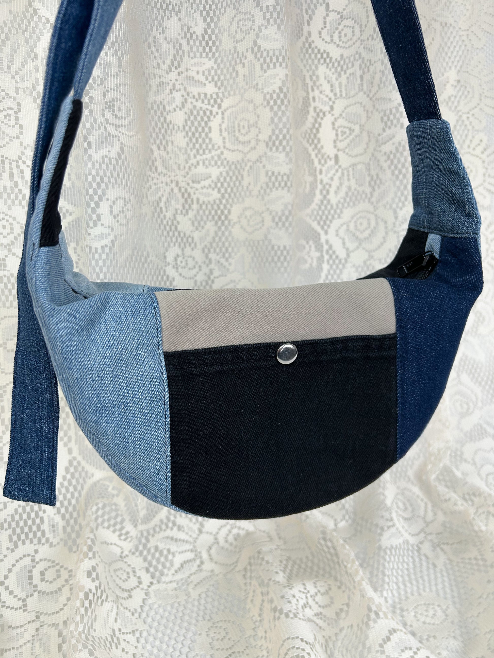Close up of the multi-coloured denim crescent bag with slip pocket with a metal snap in front of a white lace backdrop