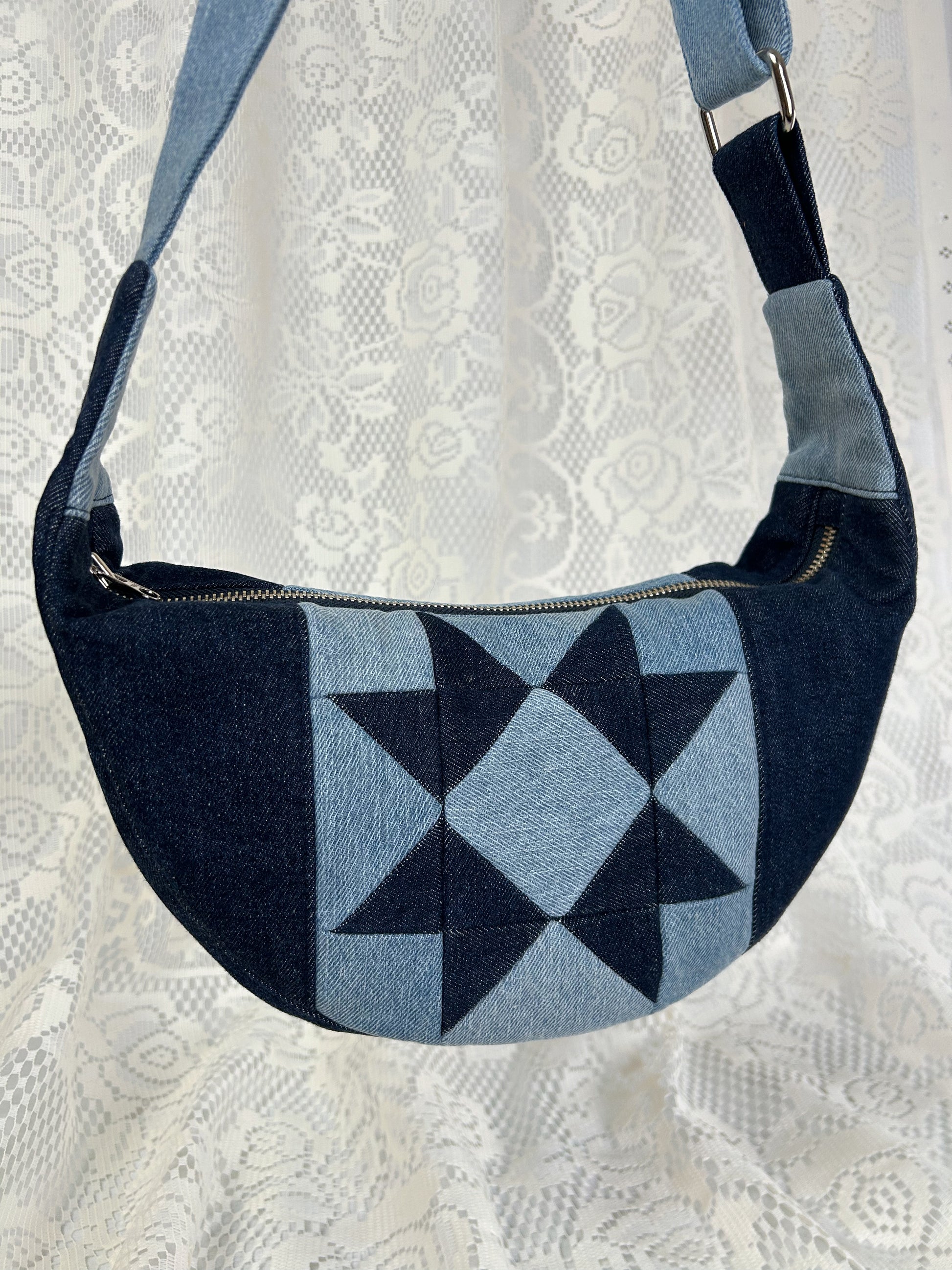 Close up of a two tone blue denim crescent bag with a patchwork star pattern in front of a white lace backdrop