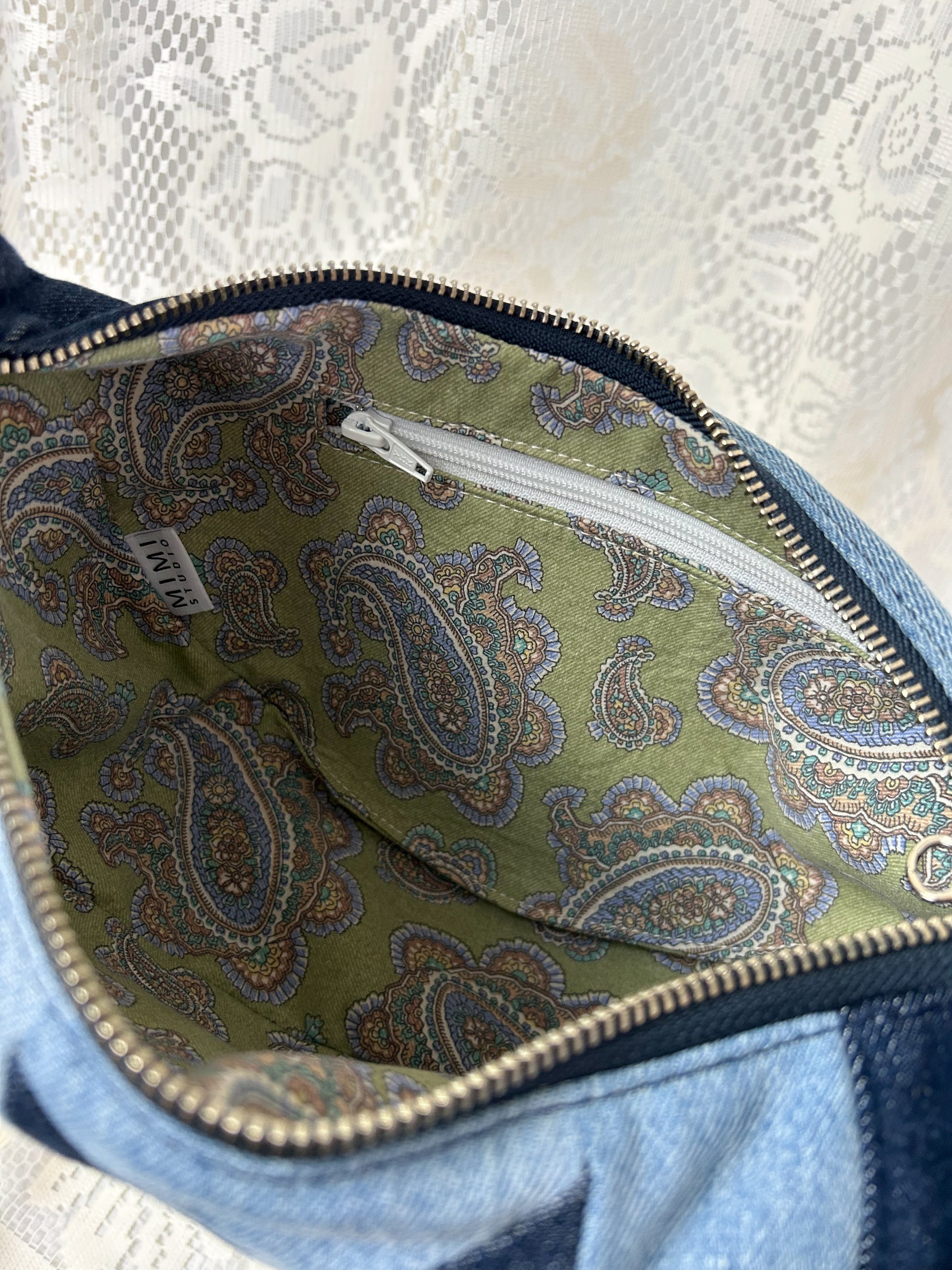 Close up of the lining fabric for the two tone blue denim crescent bag which is a paisley pattern cotton and has a zippered pocket, in front of a white lace backdrop