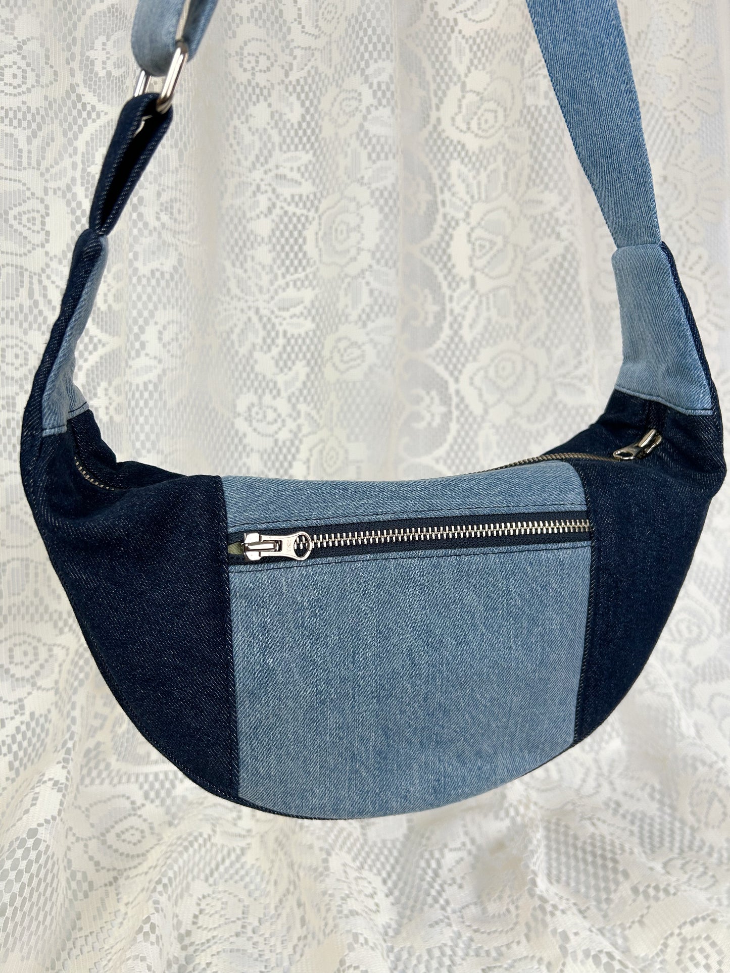 Close up of a two tone blue denim crescent bag with a zippered pocket in front of a white lace backdrop