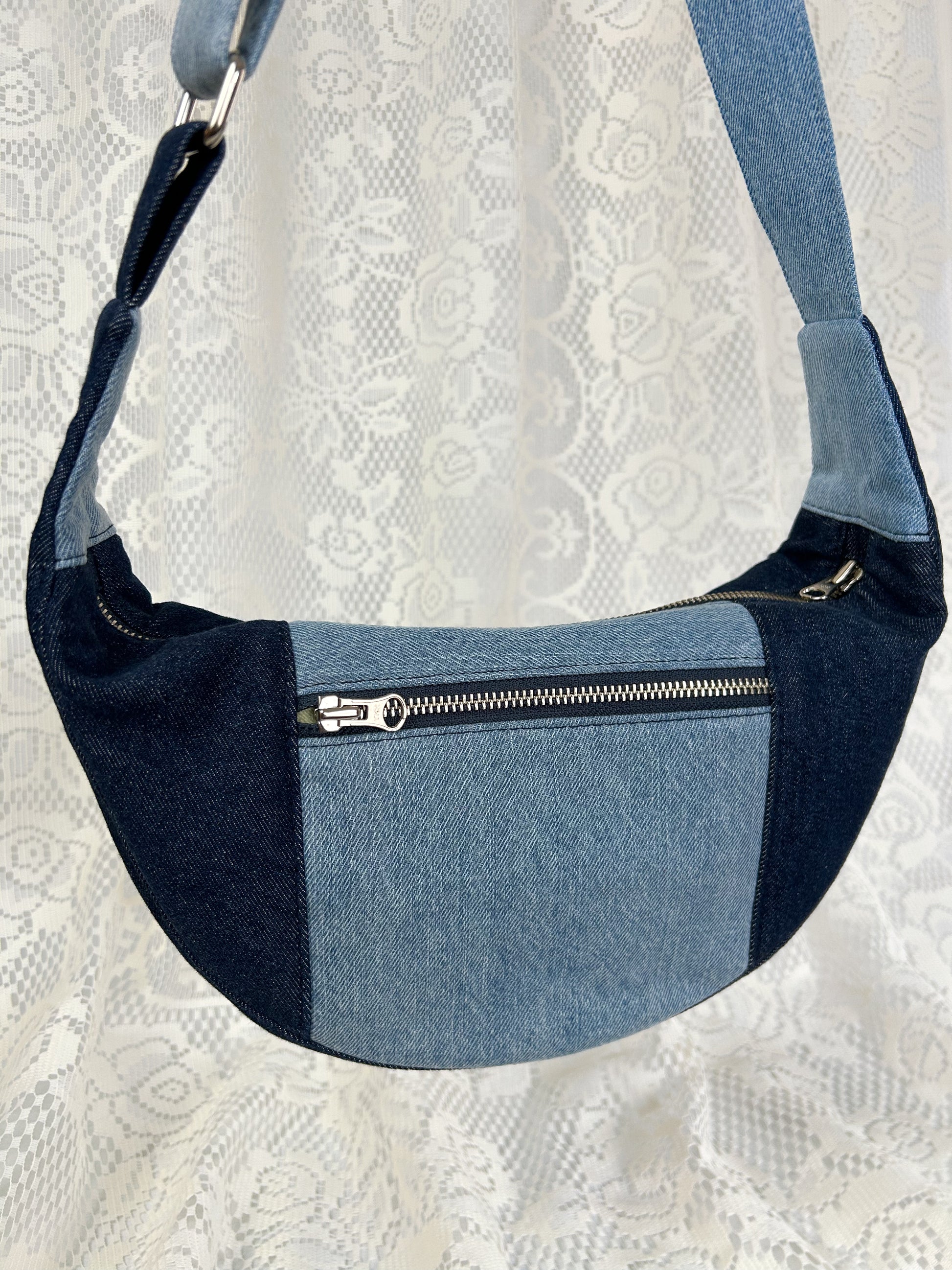 Close up of a two tone blue denim crescent bag with a zippered pocket in front of a white lace backdrop