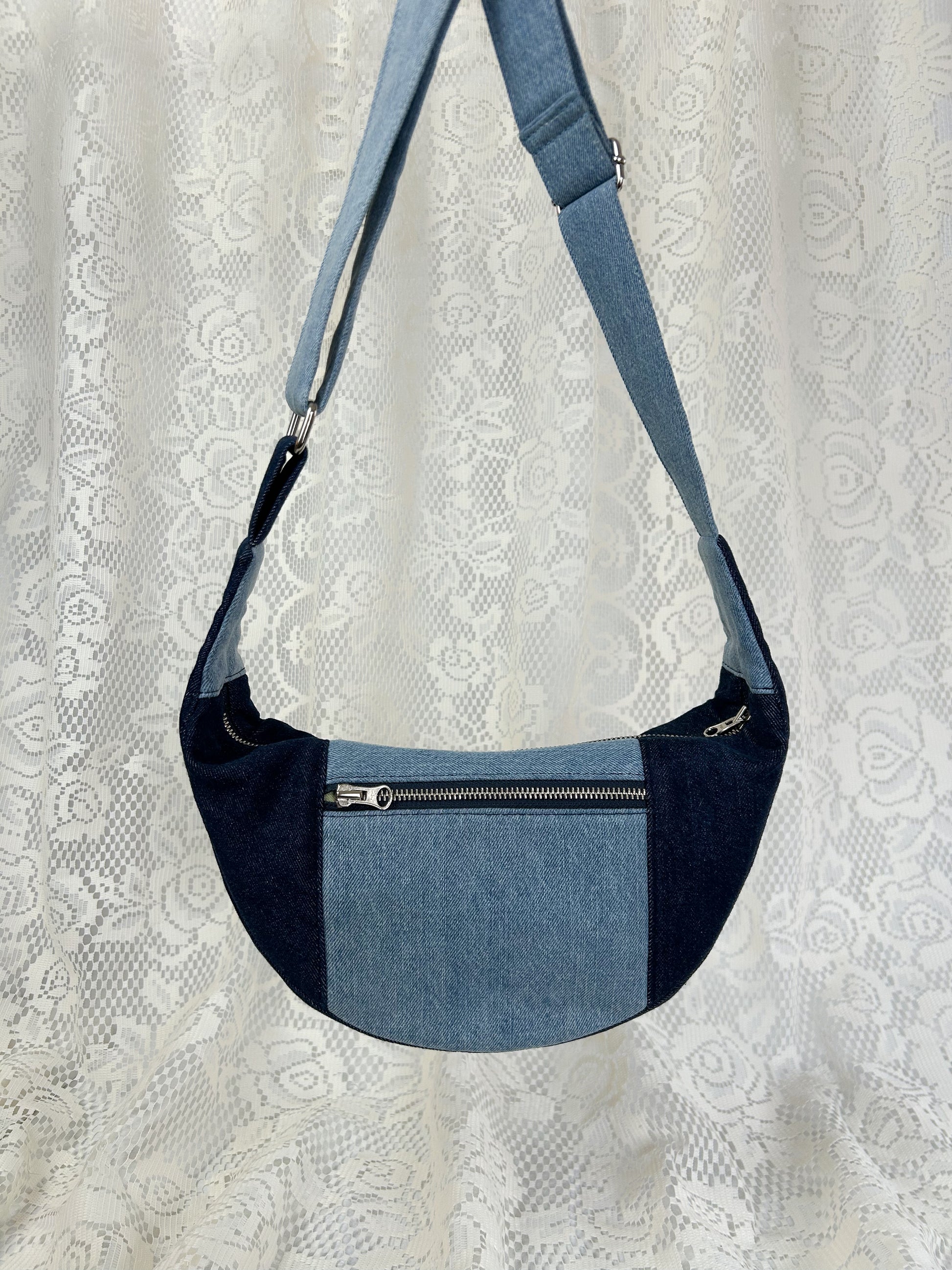 Two tone blue denim crescent bag with an exterior zippered pocket in front of a white lace backdrop