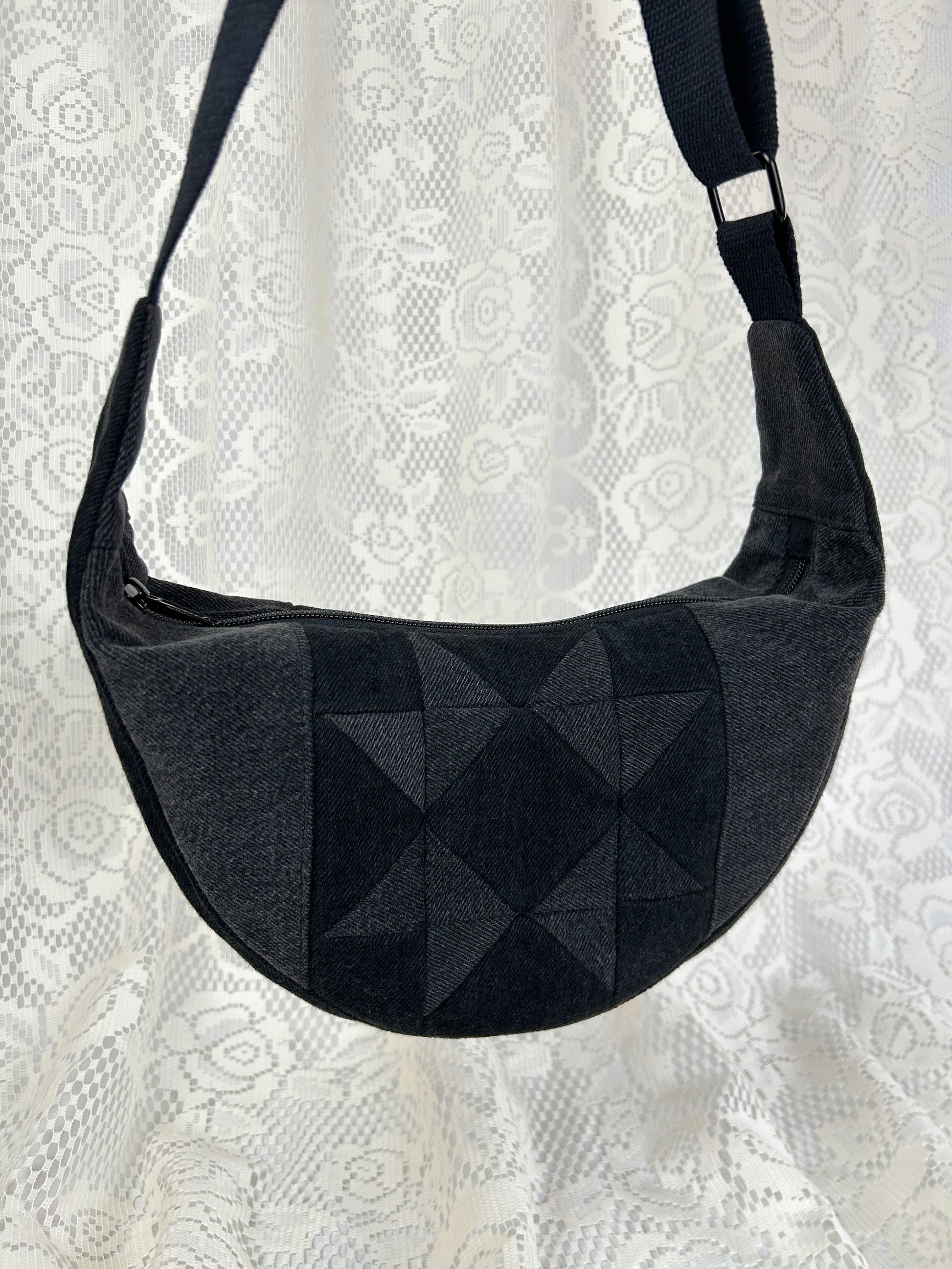 Close of up the black and grey denim crescent bag with a patchwork star pattern in front of a white lace backdrop.