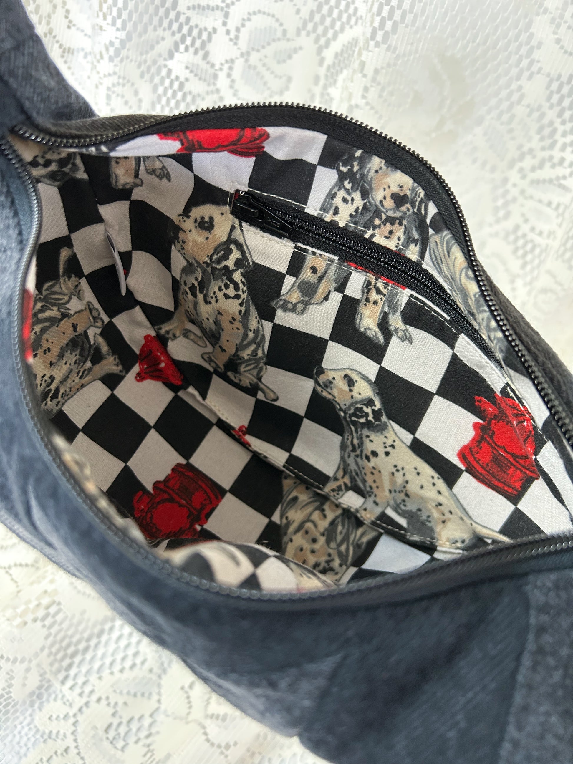 Close up of the lining fabric for the black and grey denim crescent bag which is a poly-cotton blend with a dalmation and fire hydrant pattern and has a zippered pocket, in front of a white lace backdrop
