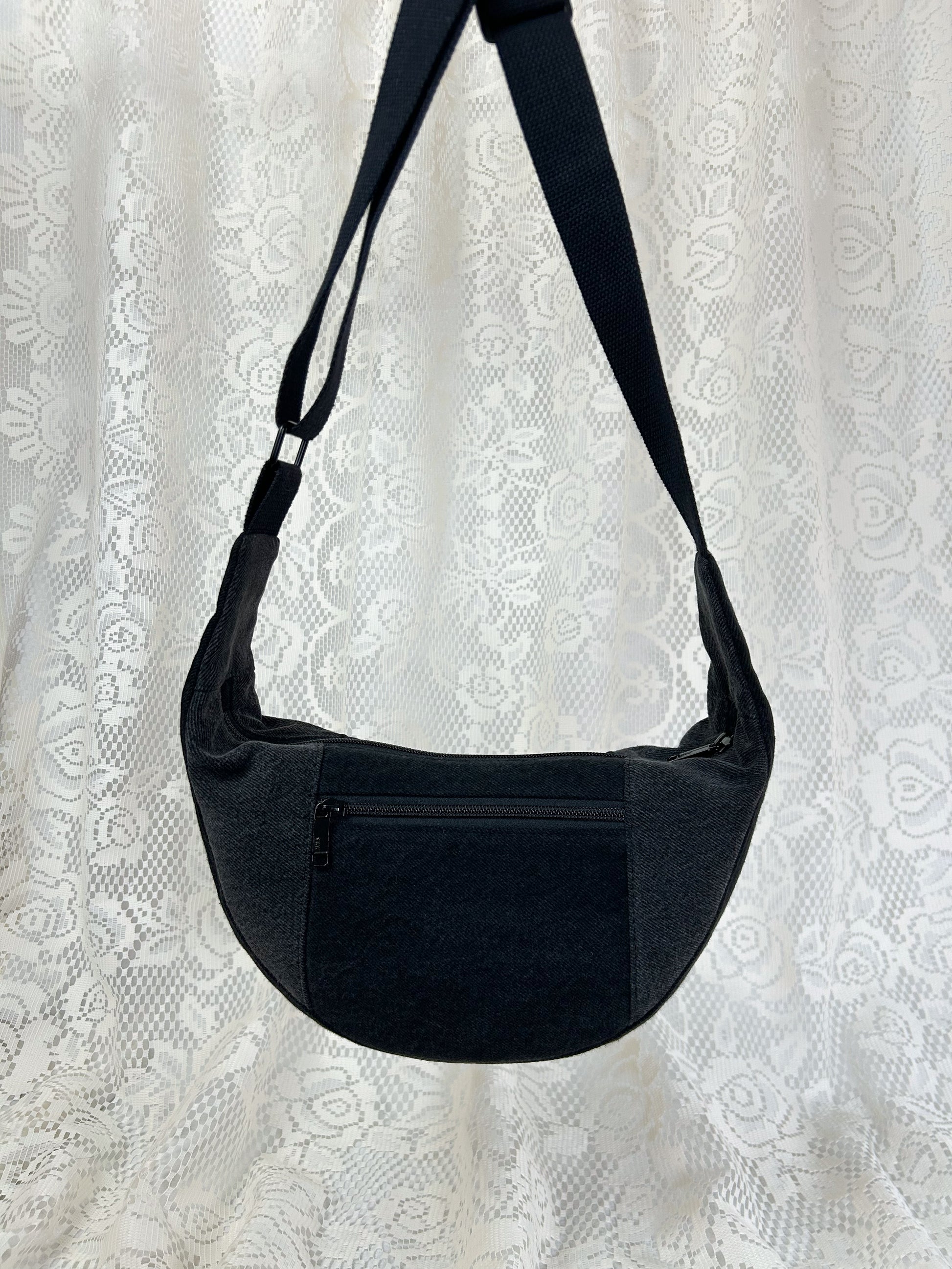 Black and grey denim crescent bag with an exterior zippered pocket in front of a white lace backdrop.