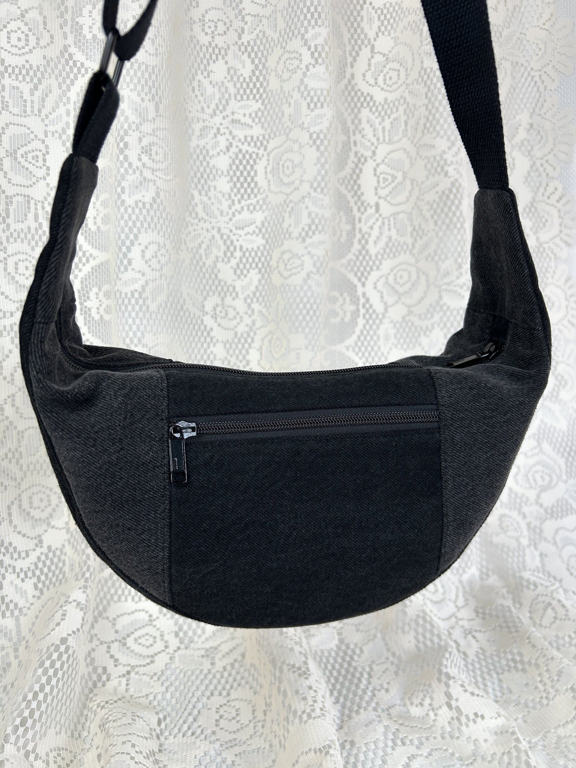 Close up of the black and grey denim crescent bag with a zippered pocket in front of a white lace backdrop.