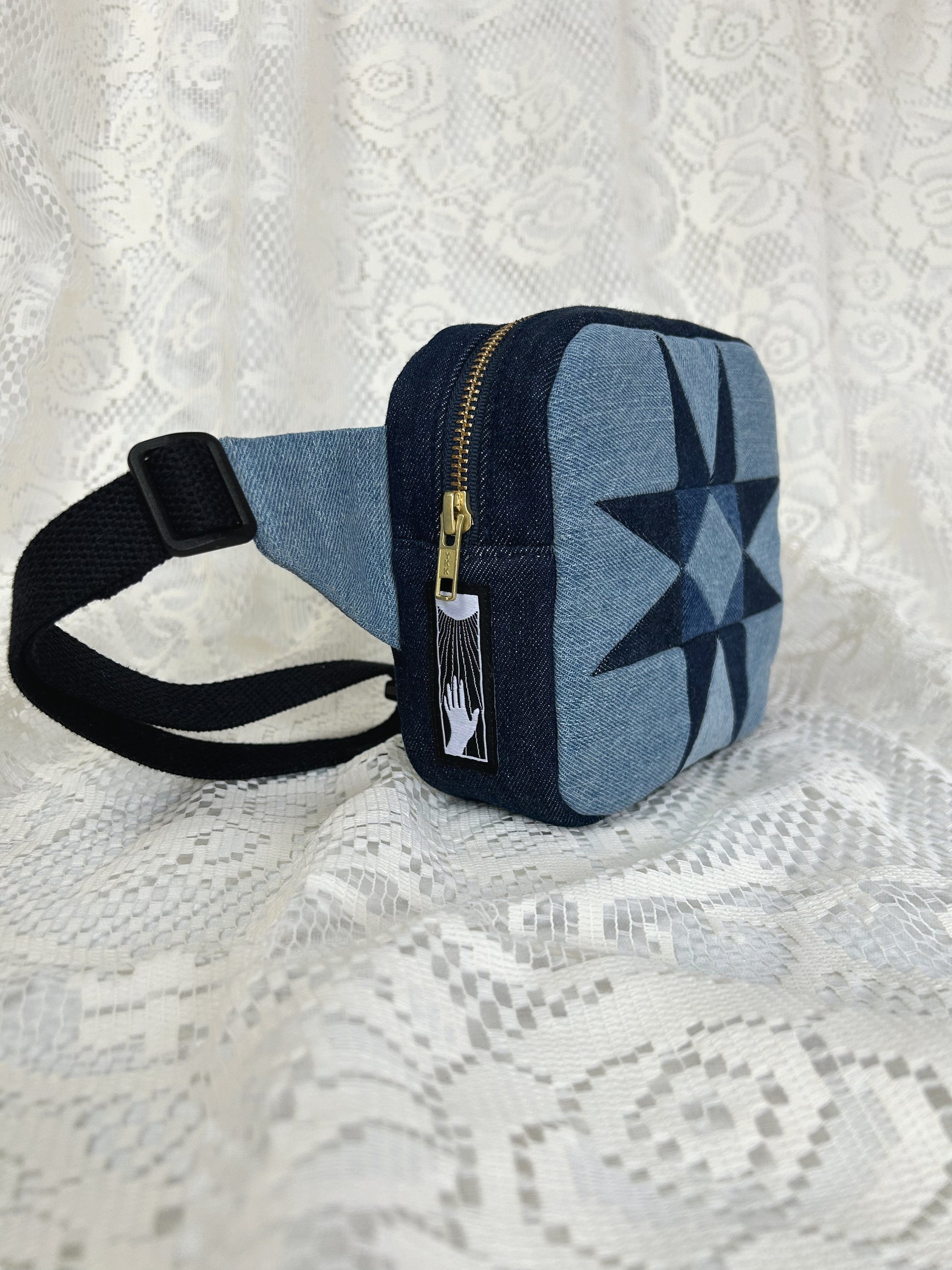 Side view of the two tone blue denim fanny pack with a patchwork star pattern showing the brass zipper and strap on a white lace background