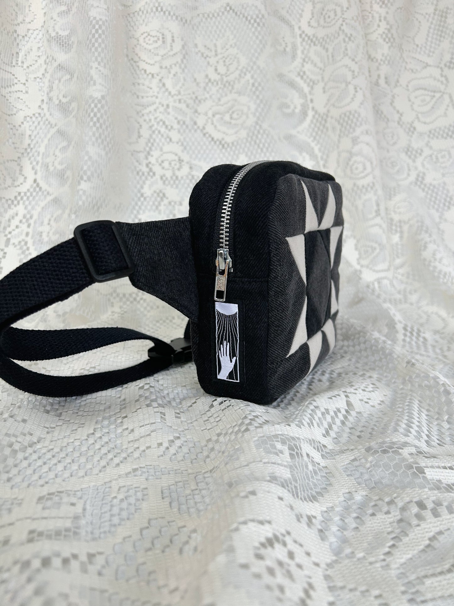 Side view of the grey, black and ivory denim fanny pack with a patchwork star pattern showing the aluminum zipper and strap on a white lace background