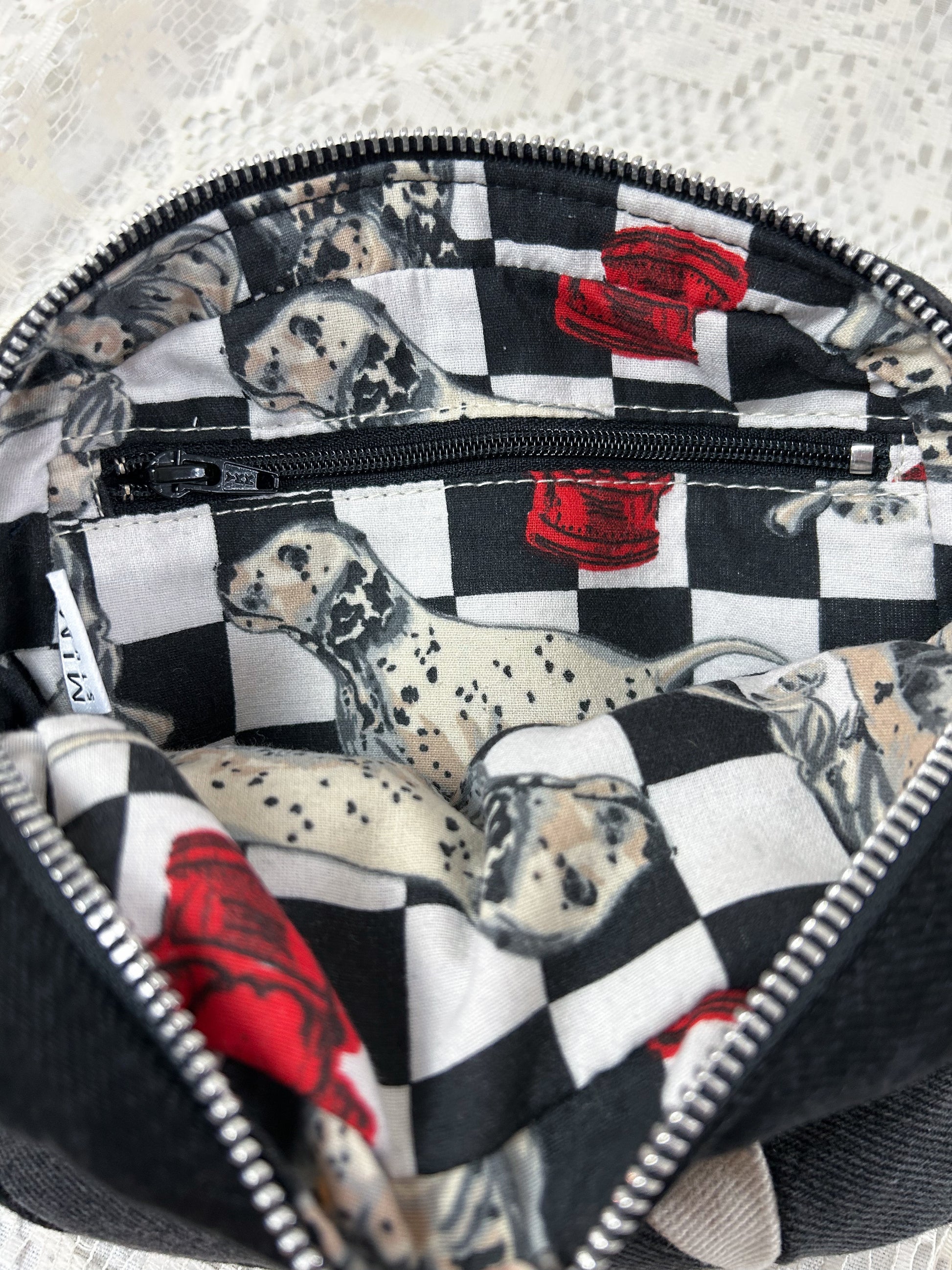 Lining fabric for the grey, black and ivory denim fanny pack with a patchwork star pattern which is a cotton-poly blend fabric with a dalmation and fire hydrant pattern and has a zippered pocket, on a white lace background