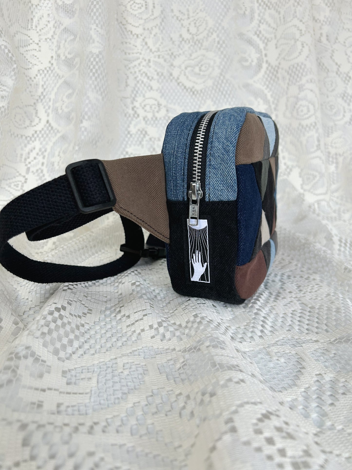 Side view of the multi-coloured denim fanny pack with a patchwork star pattern showing the aluminum zipper and strap on a white lace background