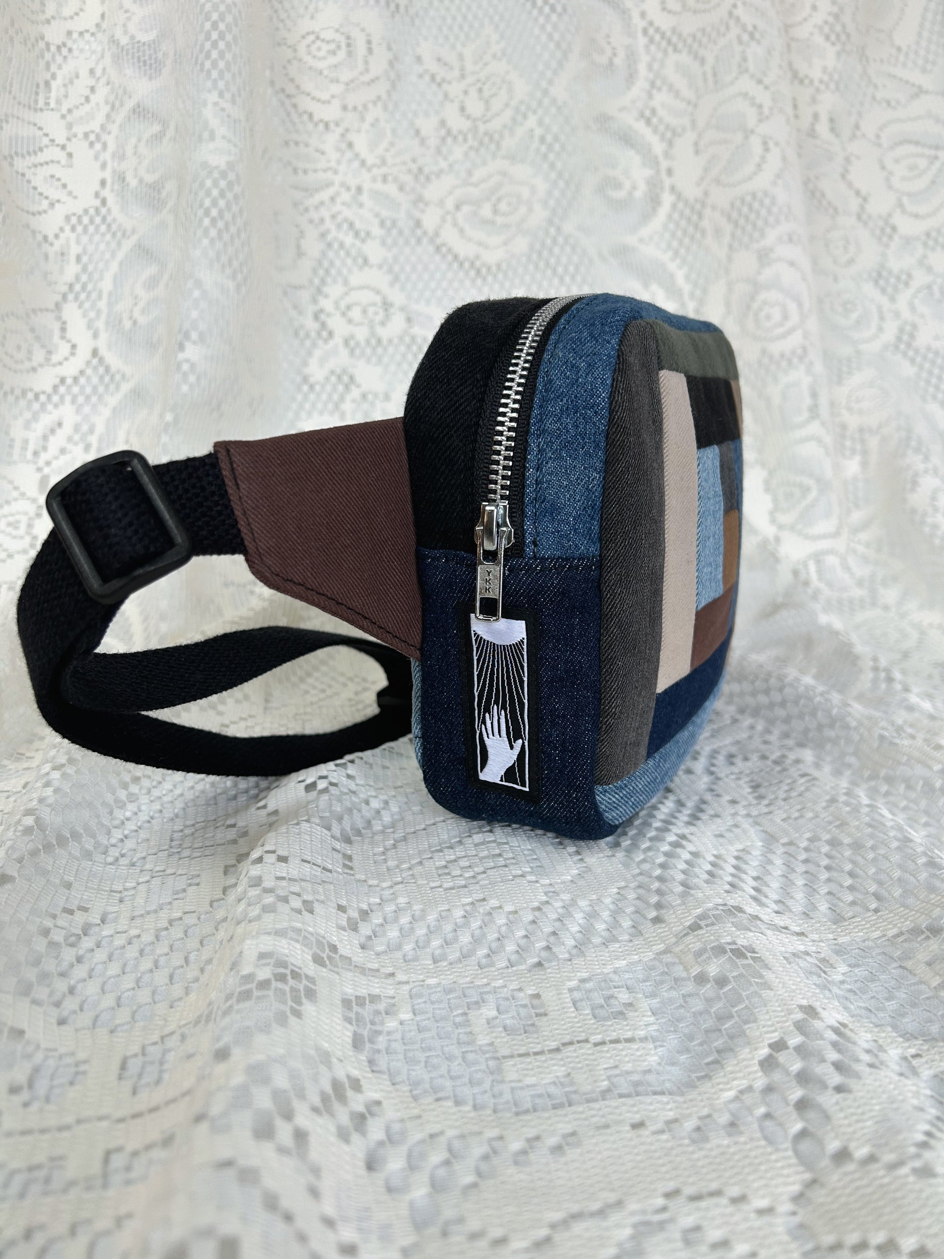 Side view of the log cabin patchwork fanny pack showing the aluminum zipper and strap on a white lace background