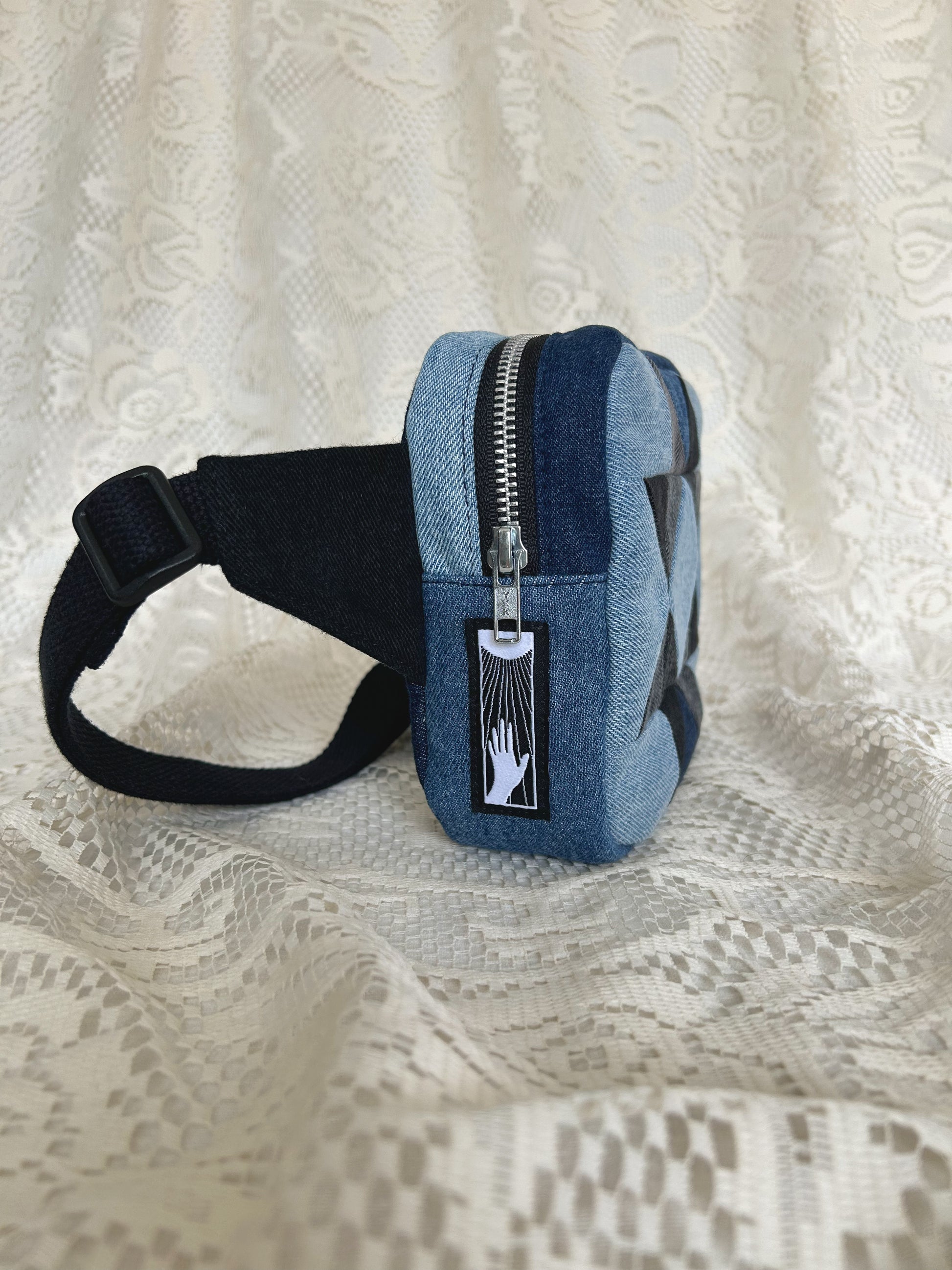 Side view of the same fanny pack showing the aluminum zipper and black cotton webbing strap with an ivory lace backdrop