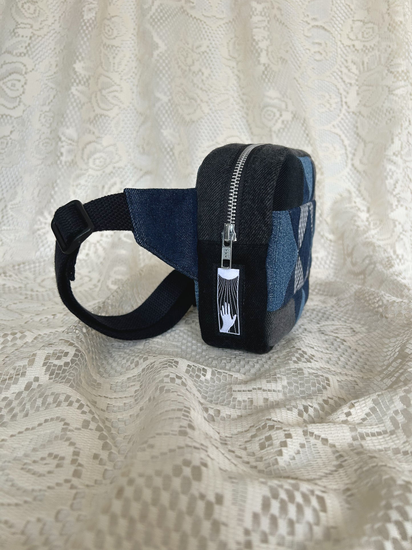 Side view of the same fanny pack showing the aluminum zipper and black cotton webbing strap with an ivory lace backdrop