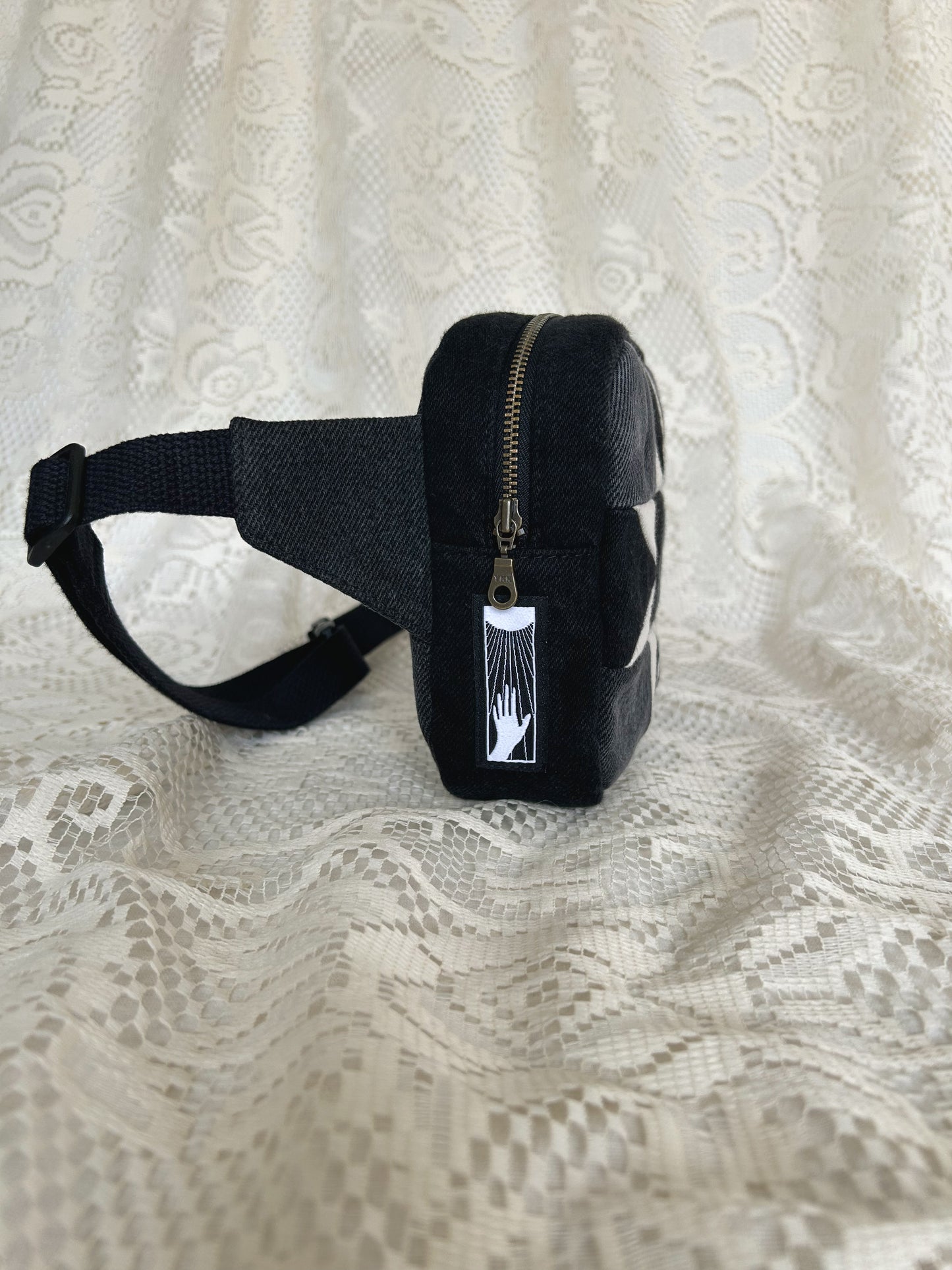 Side view of the same fanny pack showing the antique brass zipper and black cotton webbing strap with an ivory lace backdrop