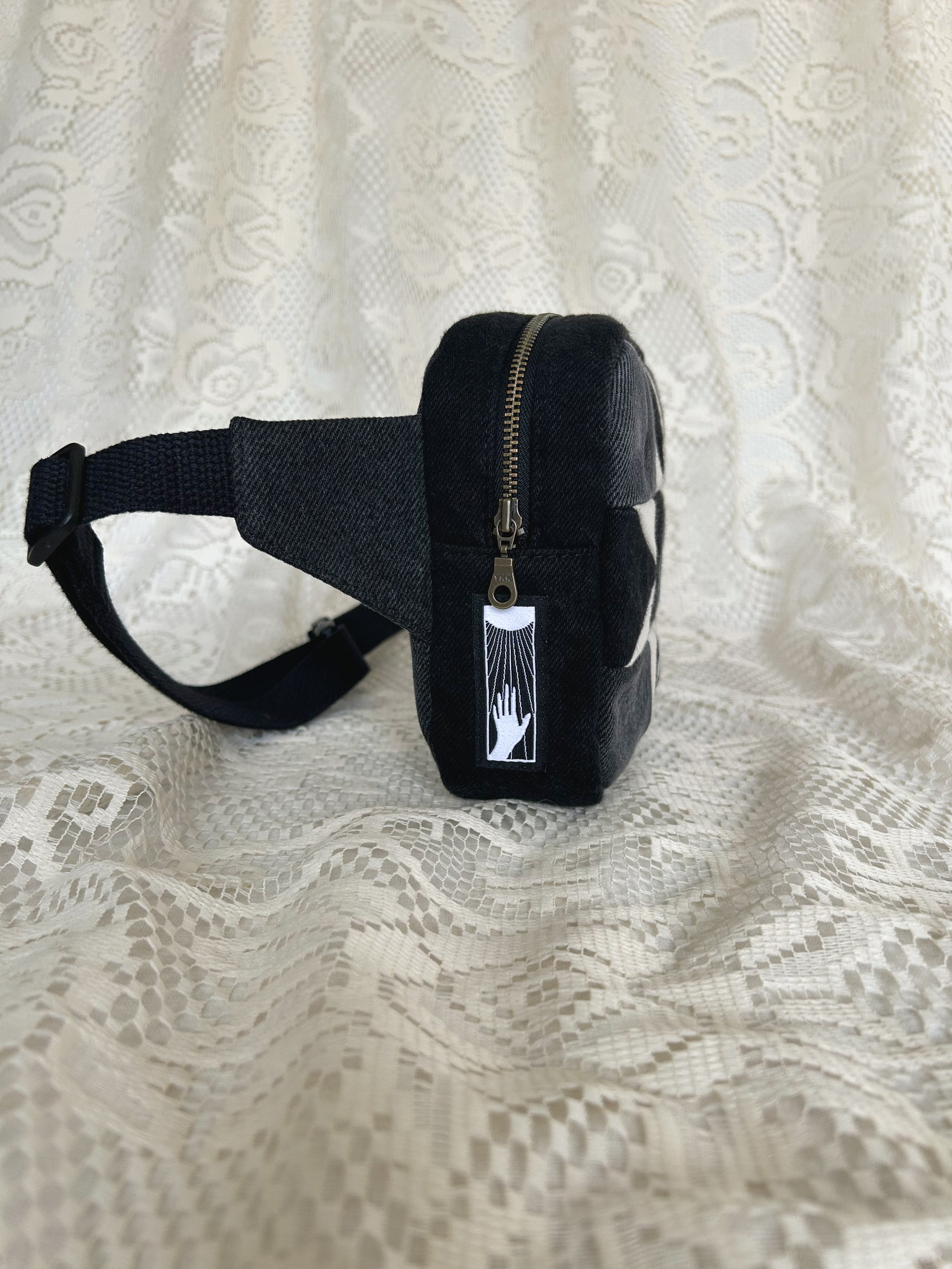 Side view of the same fanny pack showing the antique brass zipper and black cotton webbing strap with an ivory lace backdrop