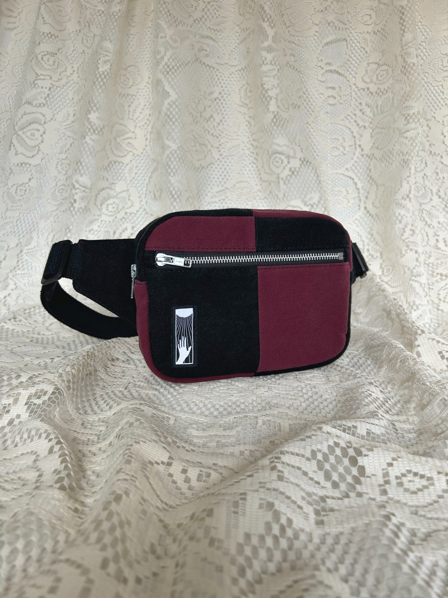 Front angled view of the half black, half burgundy denim colour block fanny pack with an ivory lace backdrop