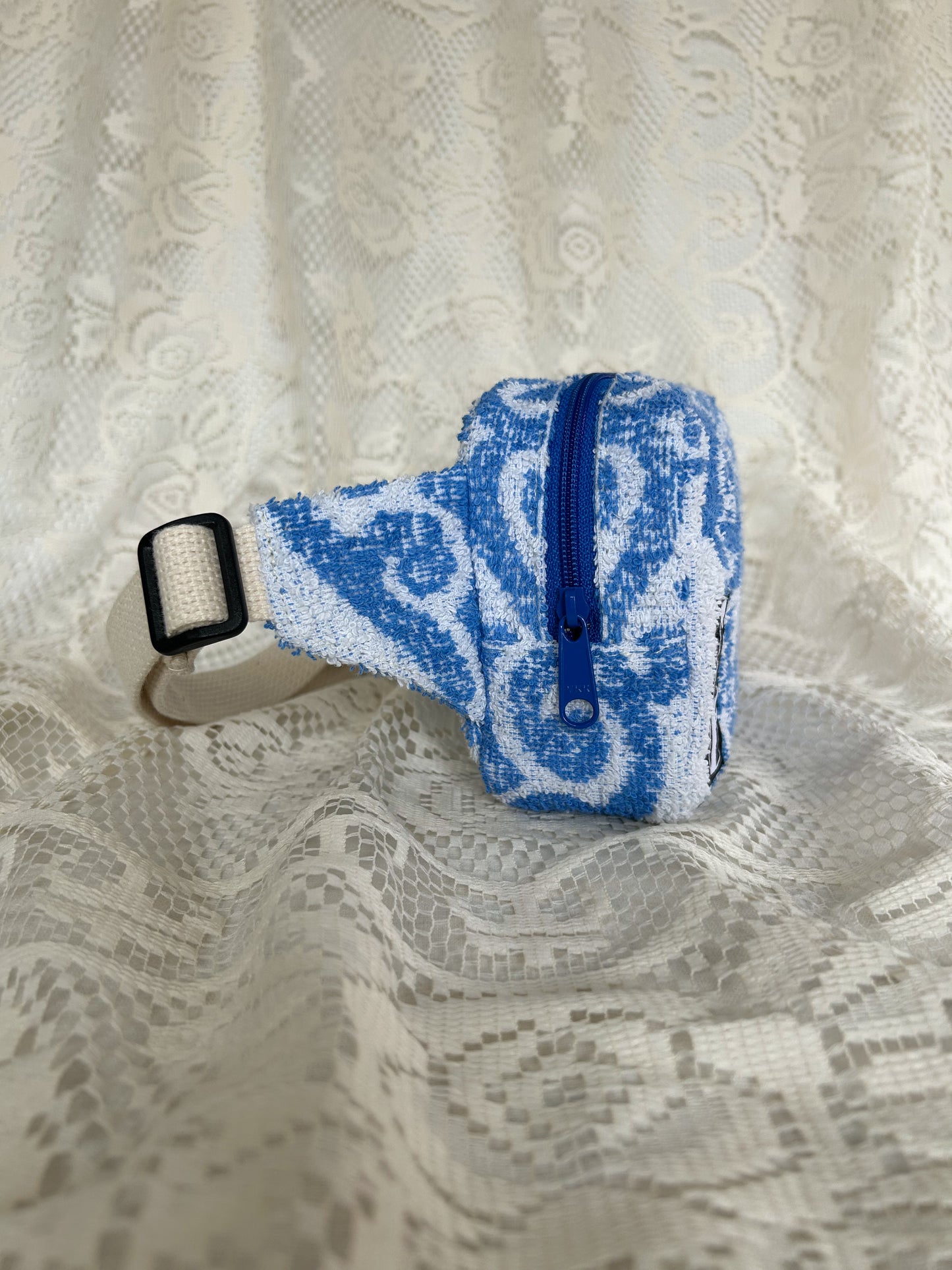 Side view of the same fanny pack showing the blue coil zipper and cream cotton webbing strap with an ivory lace backdrop