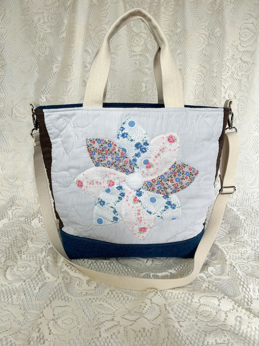 Front side of the flower quilt tote bag with a blue denim bottom and two sets of cream cotton webbing straps on an ivory lace backdrop