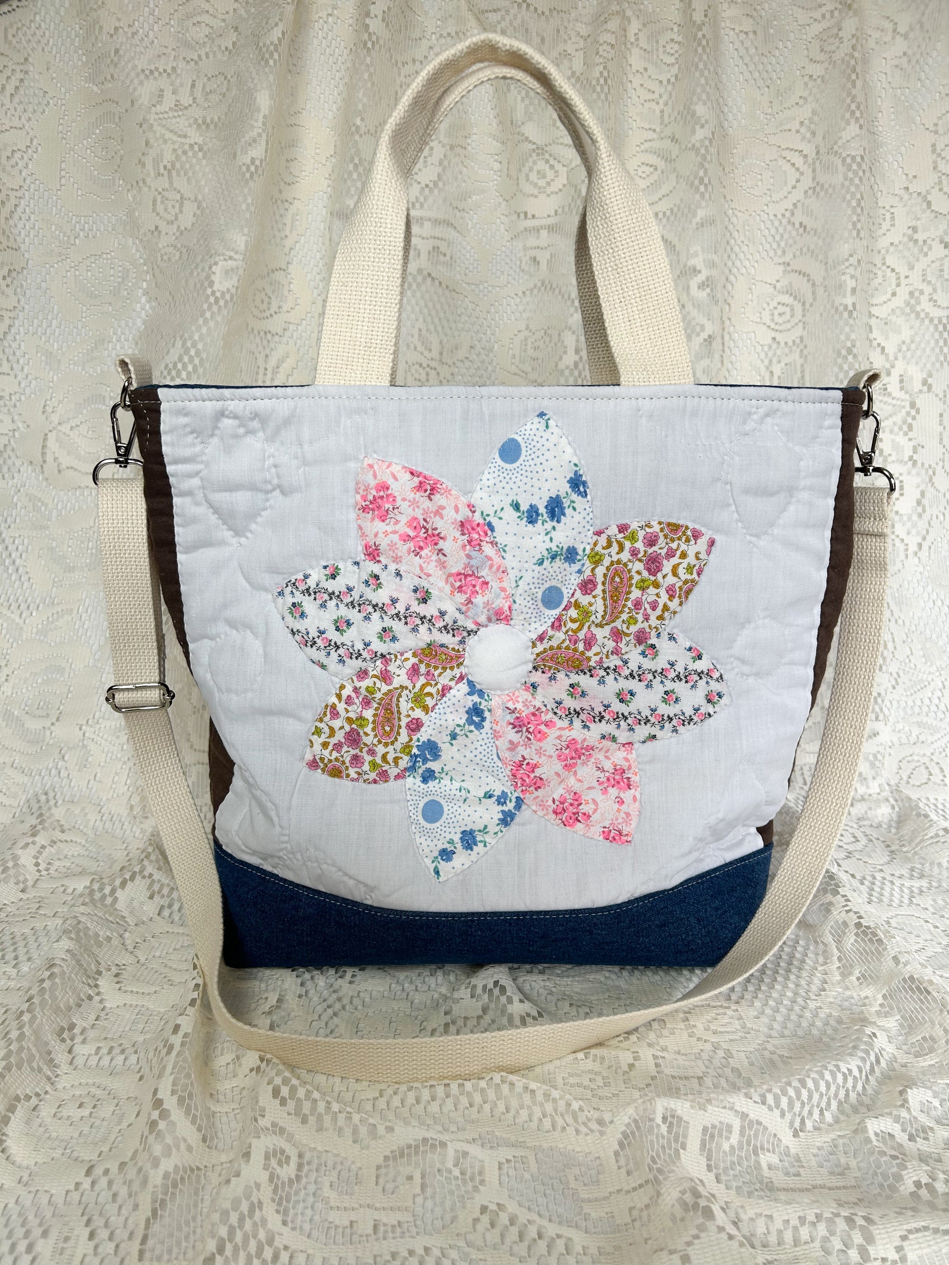 Back side of the flower quilt tote bag with a blue denim bottom and two sets of cream cotton webbing straps on an ivory lace backdrop