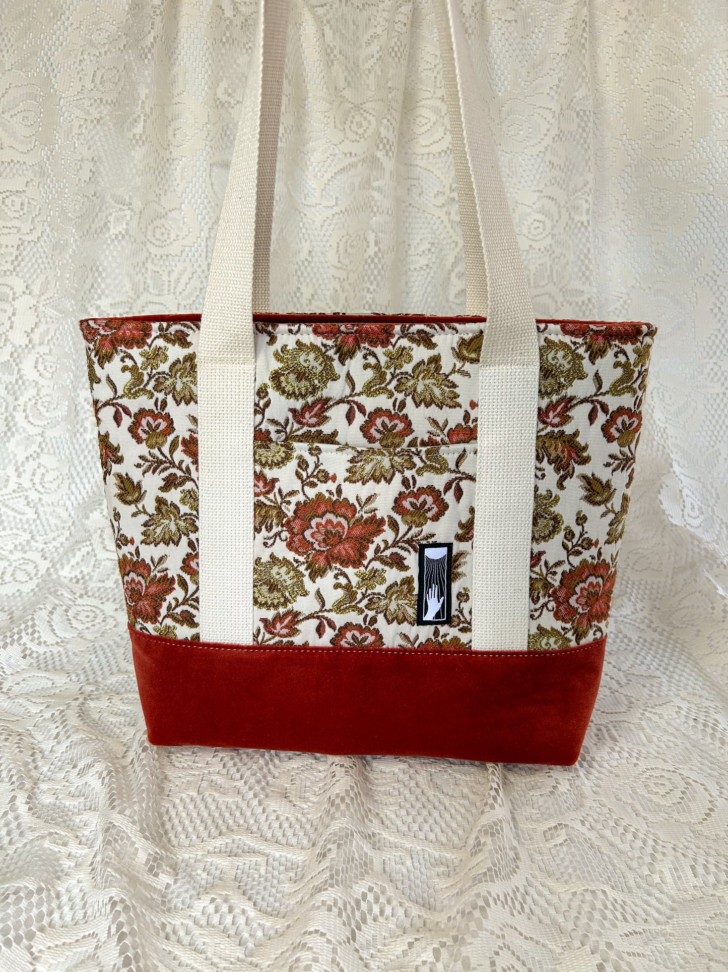A cream, rust, and gold coloured floral patterned tote bag with a rust coloured botton. The bag has cream coloured cotton webbing straps and is on an ivory lace backdrop