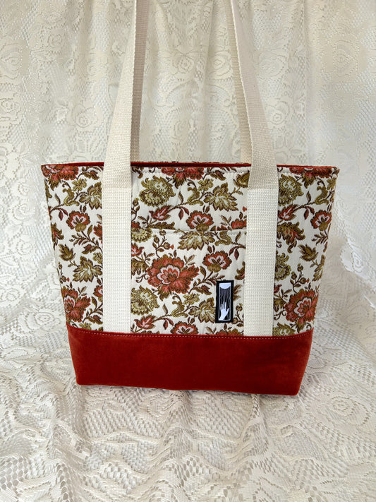 A cream, rust, and gold coloured floral patterned tote bag with a rust coloured botton. The bag has cream coloured cotton webbing straps and is on an ivory lace backdrop