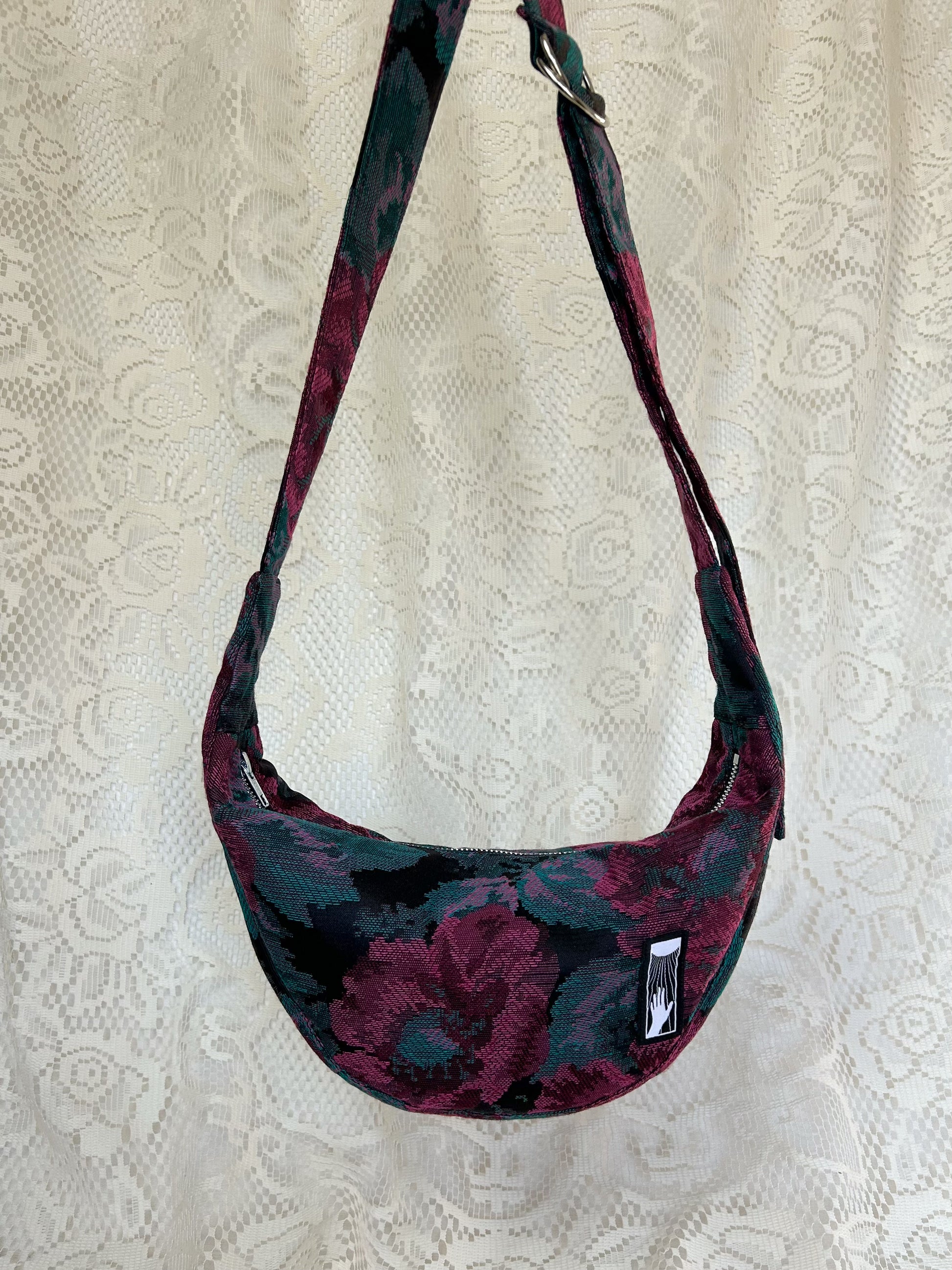 Front view of burgundy, black and turquoise floral patterned upholstery fabric crescent moon bag with an ivory lace backdrop