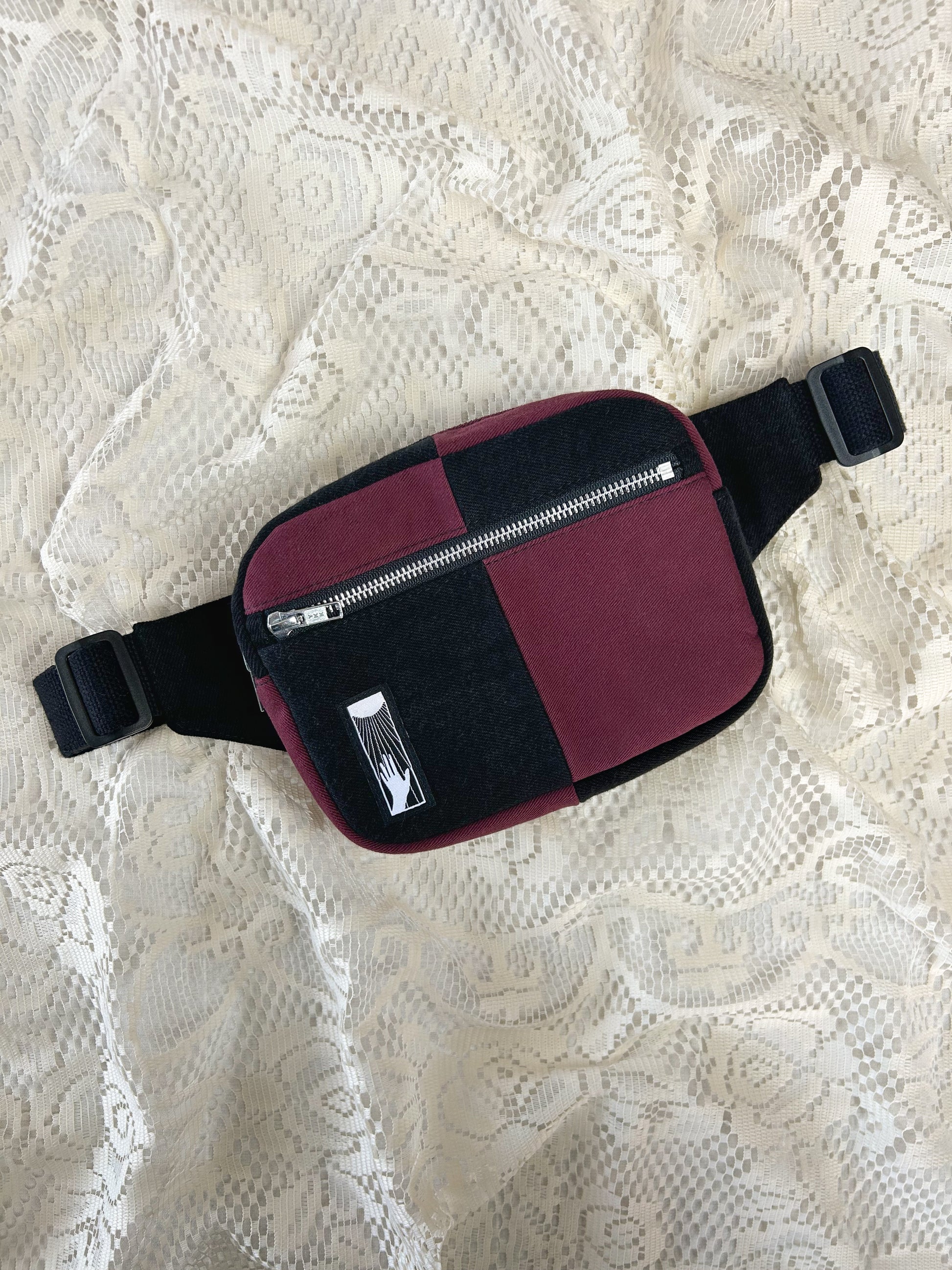Flat lay view of the half black, half burgundy denim colour block fanny pack with an ivory lace backdrop