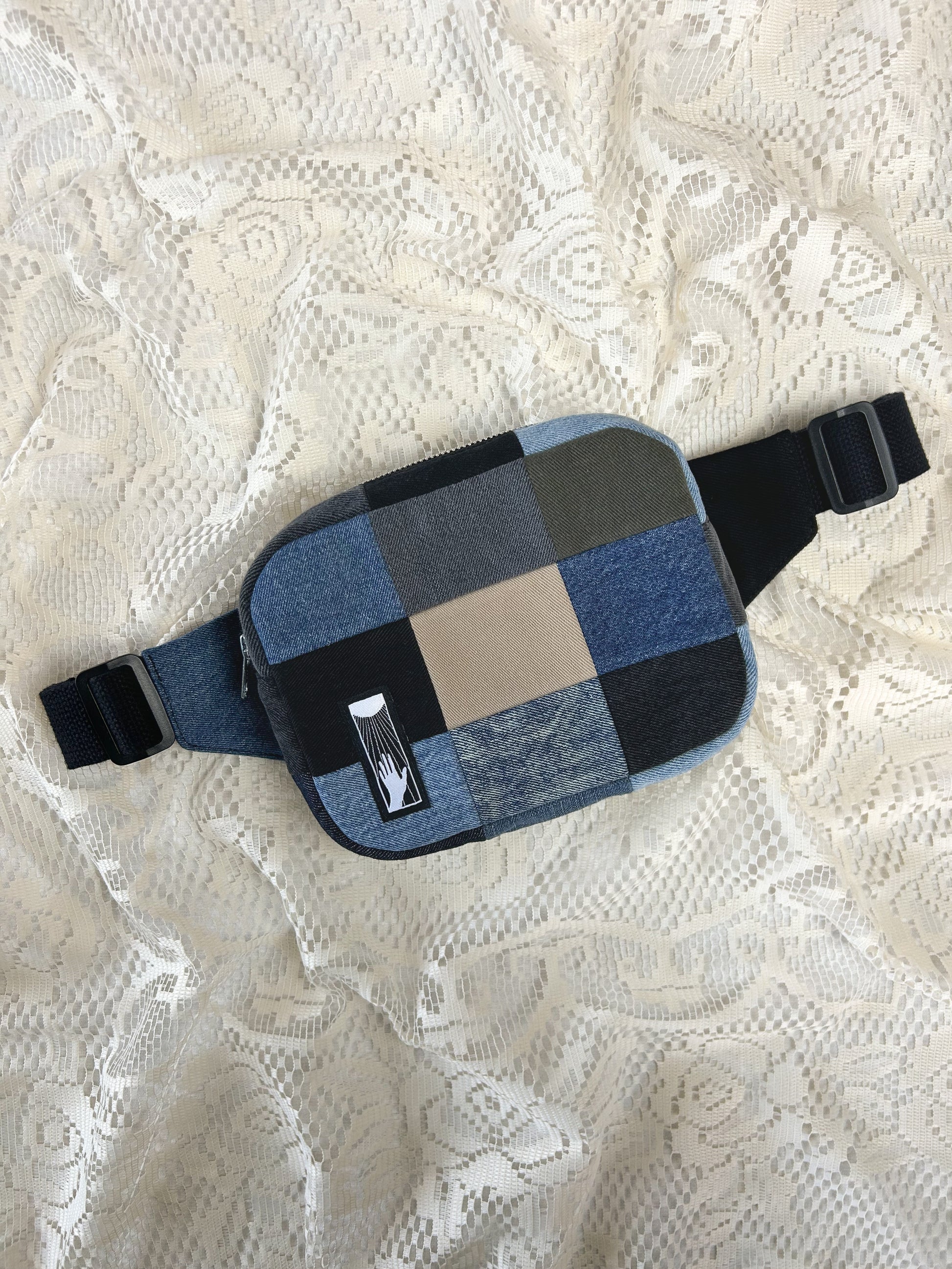 Flat lay view of a fanny pack made from various denim patches in shades of blue, ivory, black, grey and green on an ivory lace backdrop