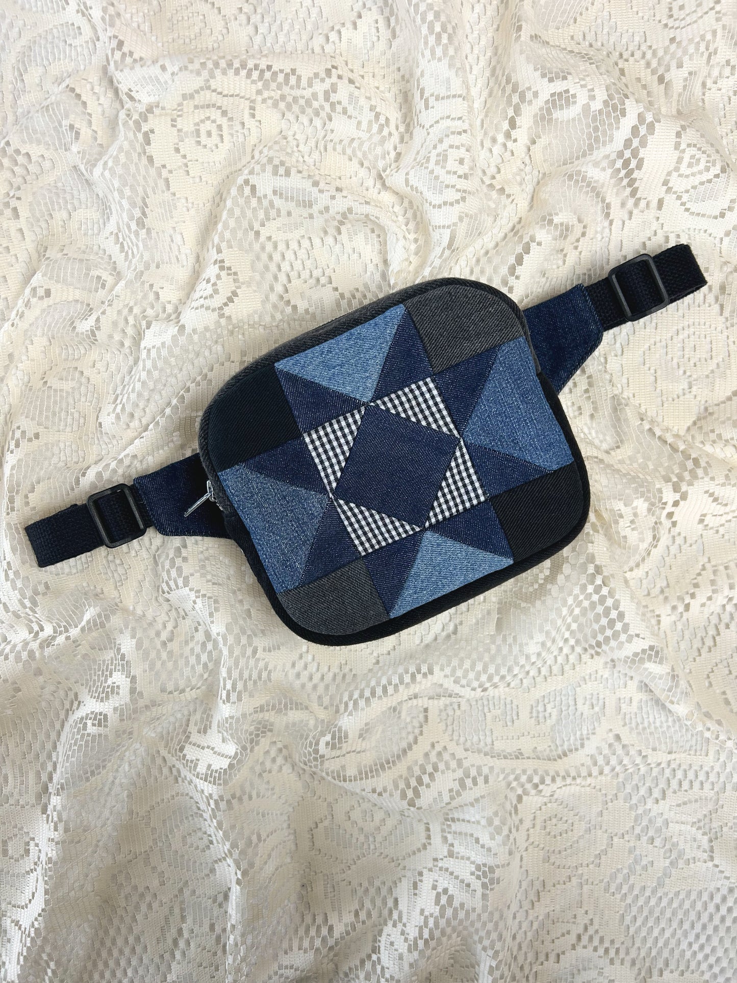 Flat lay view of a fanny pack with a star pattern made from patches of blue, grey, and black denim and gingham fabric on an ivory lace background