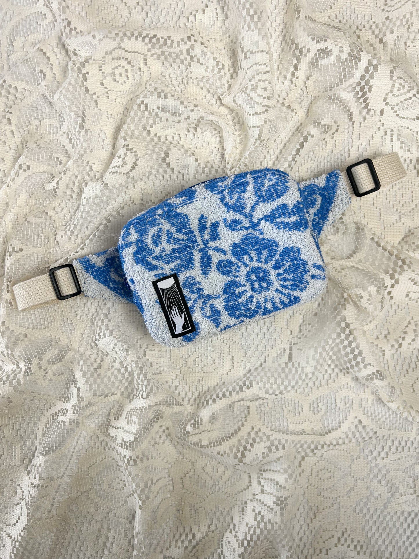 Flat lay view of a white and blue floral patterned towel fanny pack on an ivory lace background