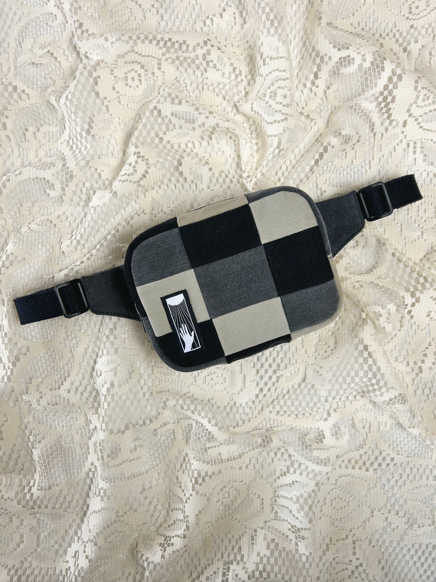 Flat lay view of a fanny pack with grey, black and ivory patches on an ivory lace background