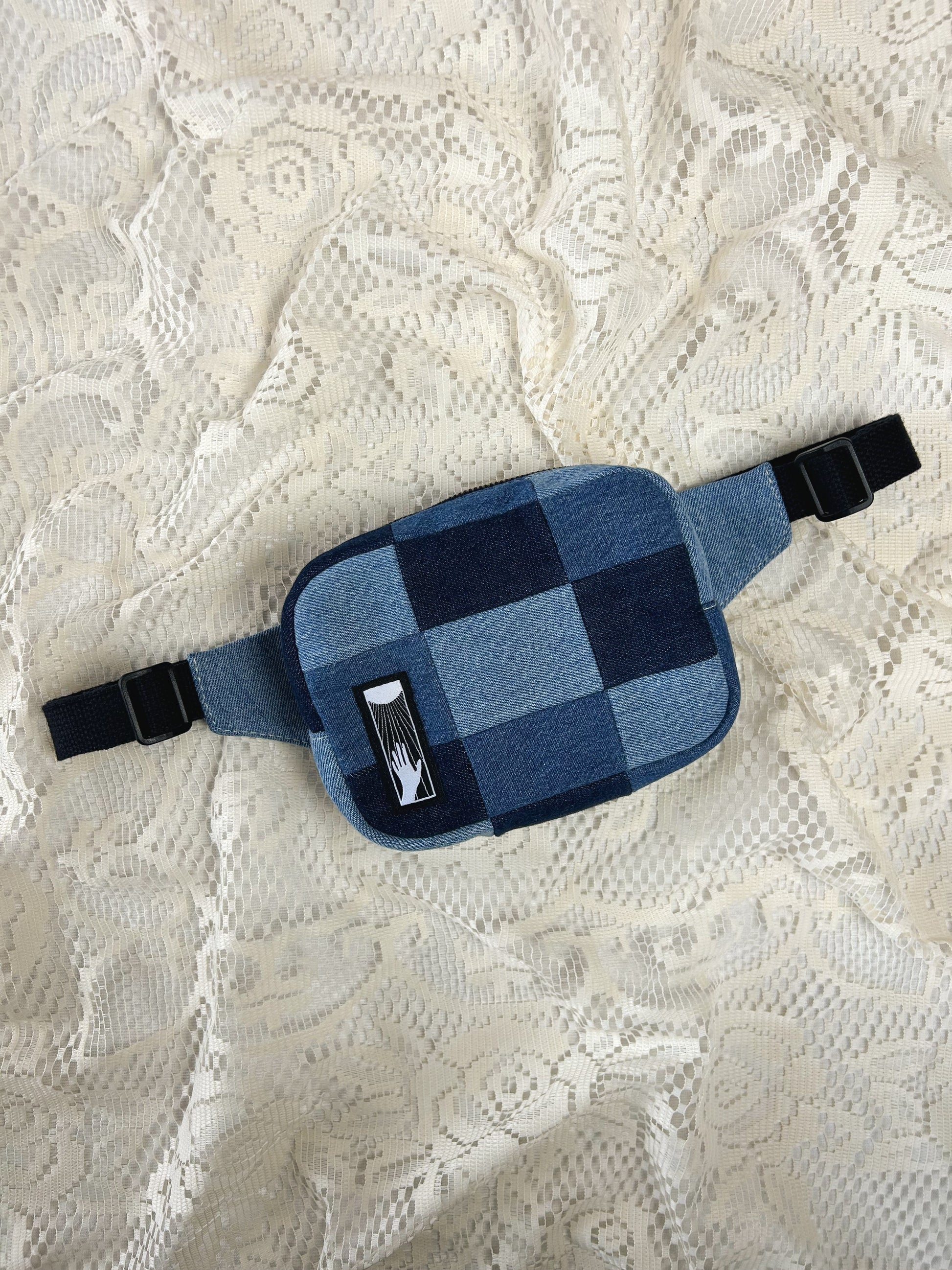 Flat lay view of a fanny pack made from patches of denim in three different shades of blue on an ivory lace background