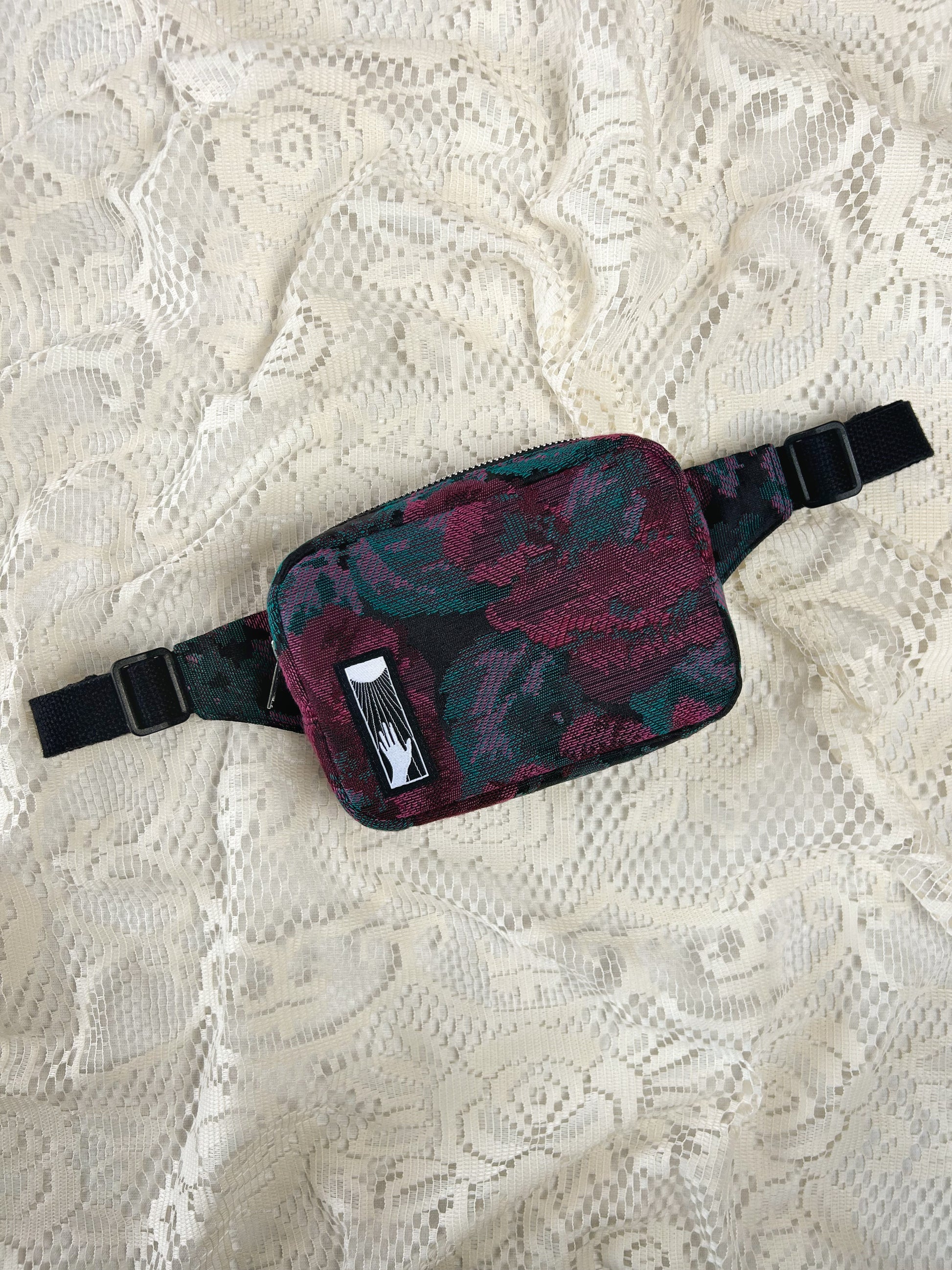 Flat lay view of a burgundy, black, and turquoise floral patterned fabric fanny pack on an ivory lace background