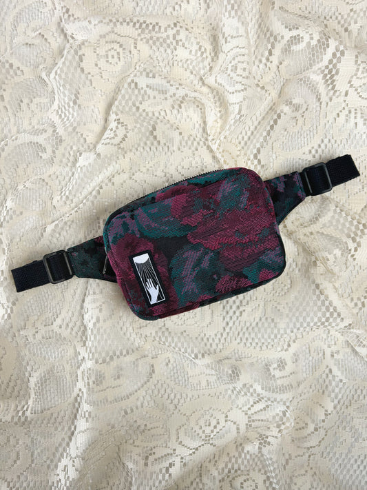 Flat lay view of a burgundy, black, and turquoise floral patterned fabric fanny pack on an ivory lace background