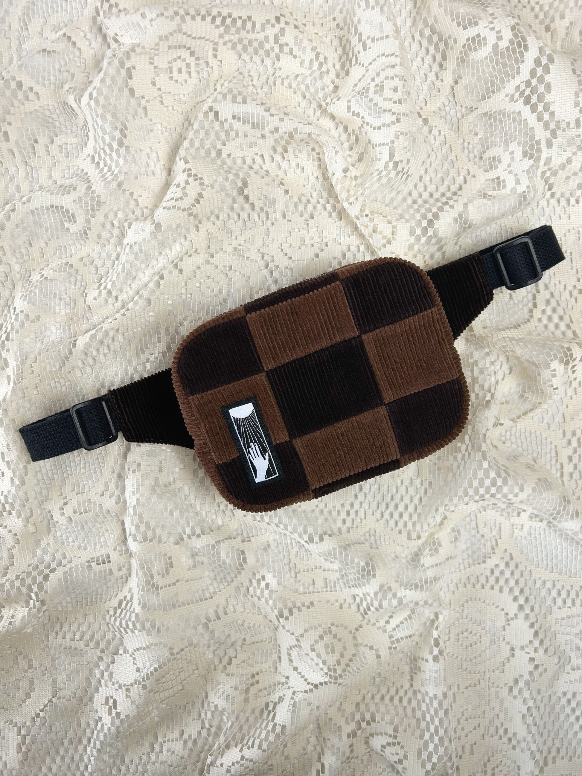 Flat lay view of a fanny pack made from patches of corduroy in 2 shades of brown on an ivory lace background