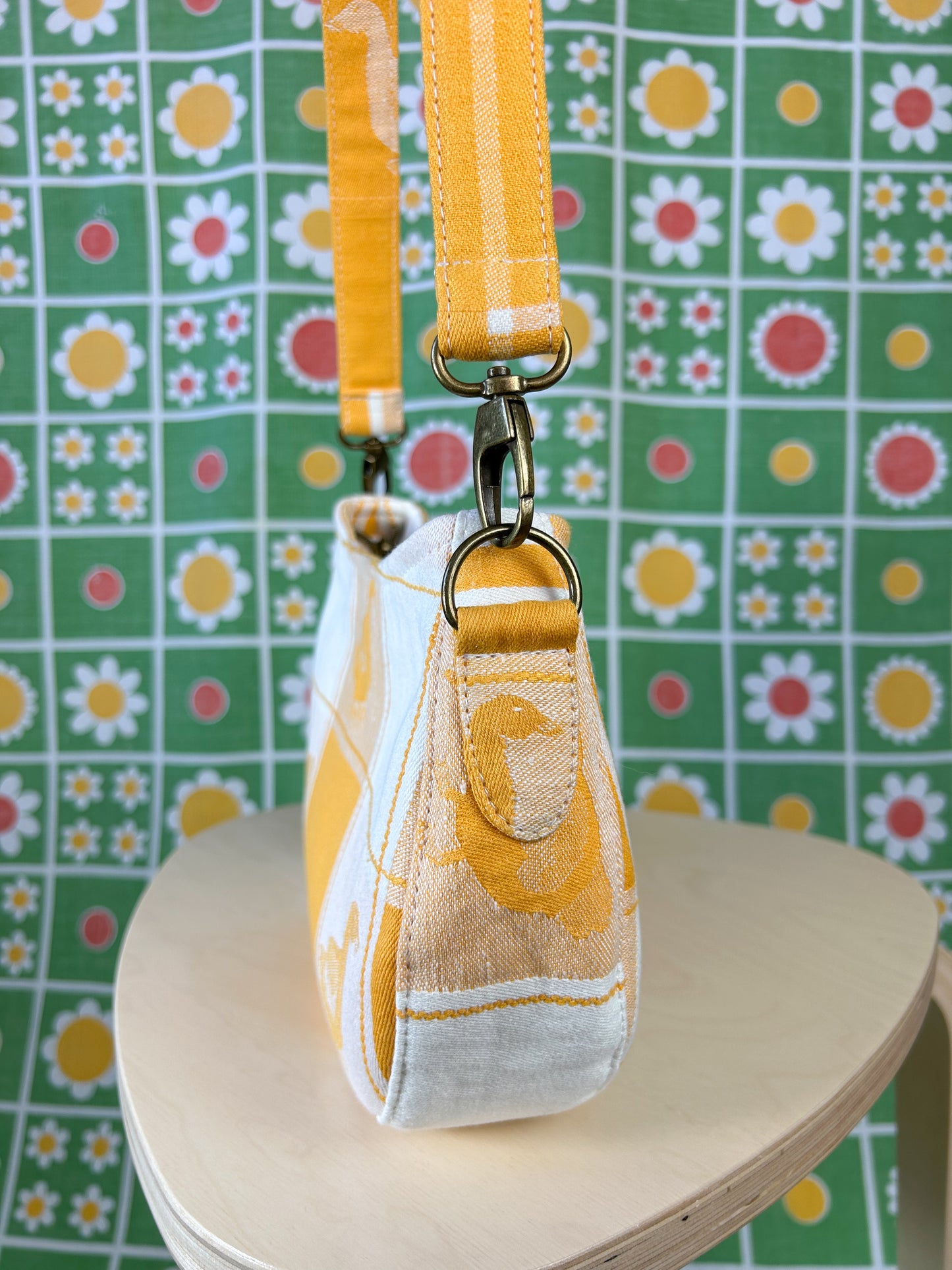 Side view of the same handbag showing the purse strap tabs, antique brass hardware and strap on a wooden stool in front of a floral backdrop