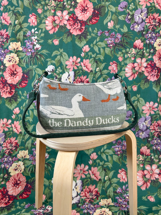 Handbag with a white duck print and  'The Dandy Ducks' text on a wooden stool against a floral background