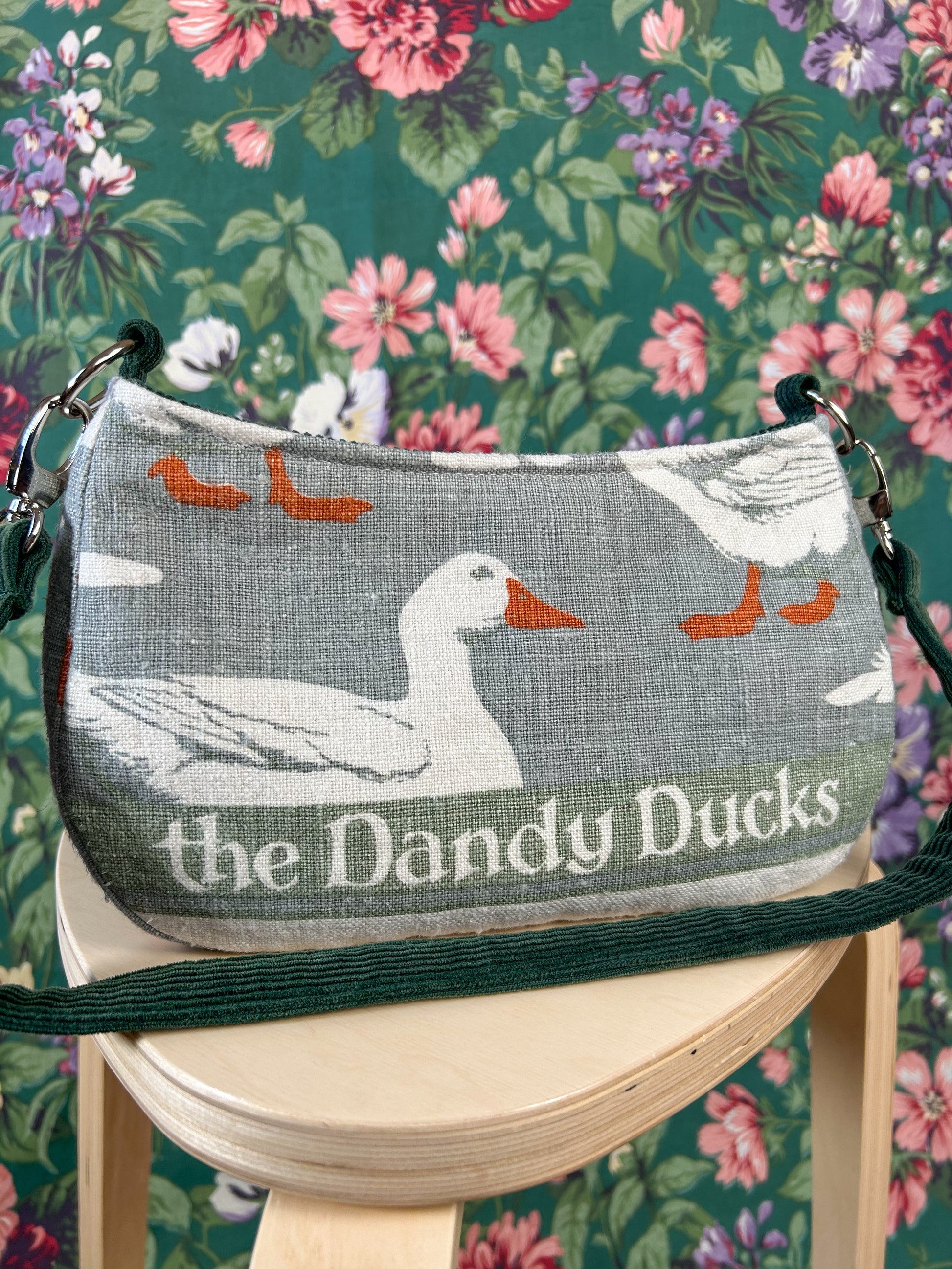 Closeup of the handbag with a white duck print and 'The Dandy Ducks' text on a wooden stool against a floral background