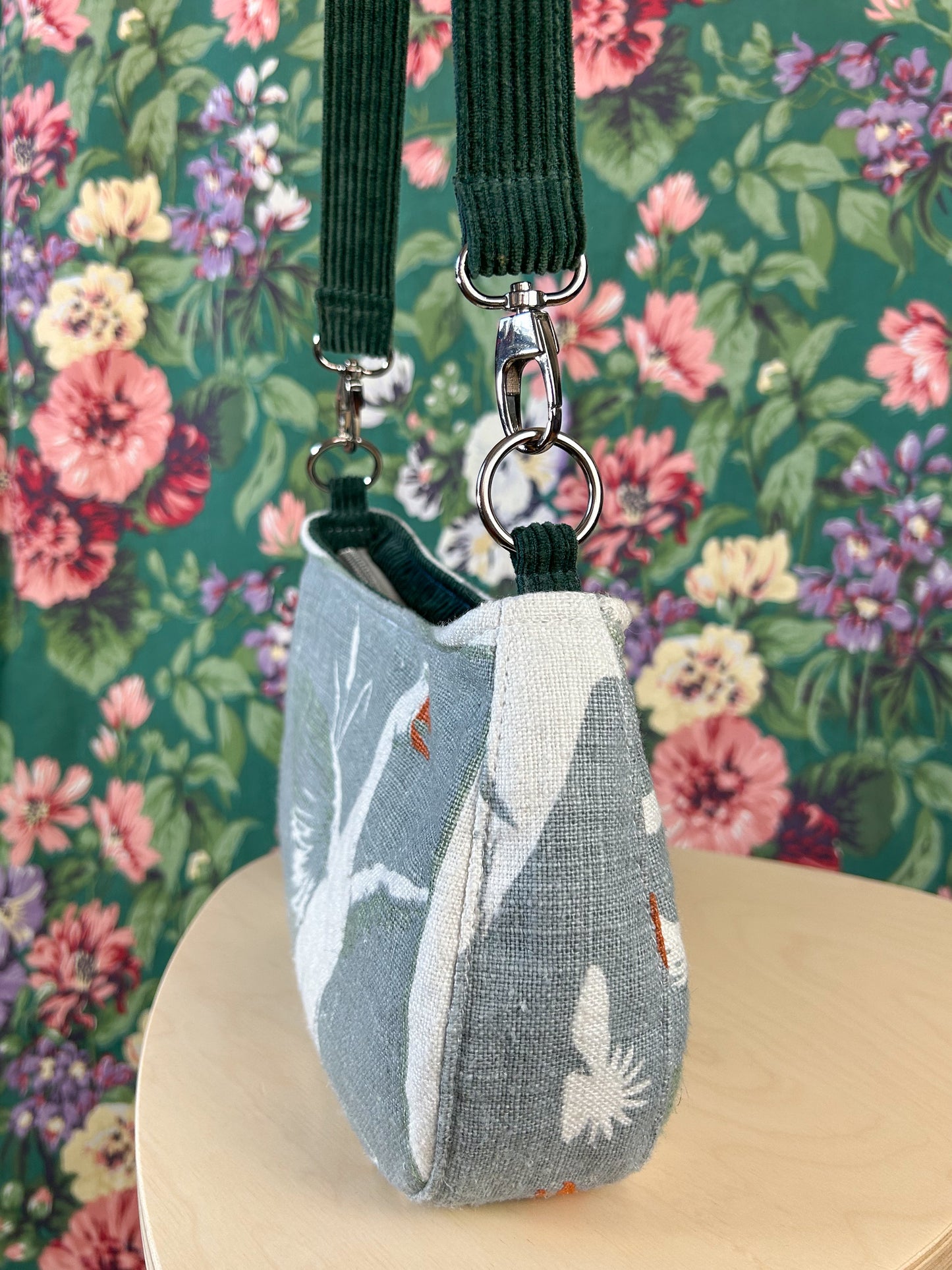 Side view of the handbag showing the strap and silver nickel hardware on a wooden stool in front of a floral backdrop